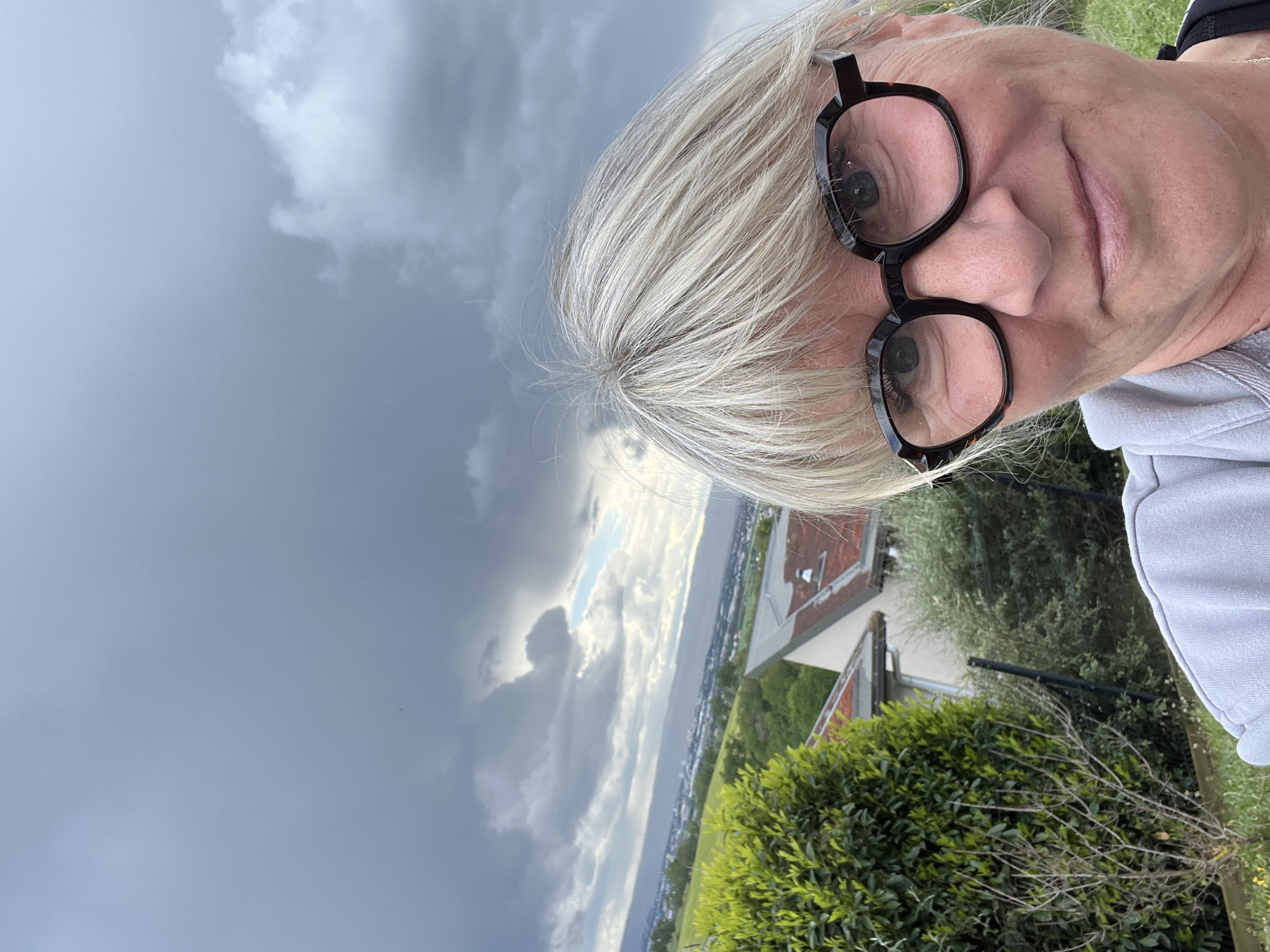 The image shows a selfie taken outdoors. A person with short, blonde hair and wearing black-rimmed glasses is prominently featured. The background displays an overcast sky, a body of water, and landscape elements. The vegetation is present, with some trees and bushes. A building is visible with a red roof. Based on visual cues, this location may be in the vicinity of Chambœuf, France.