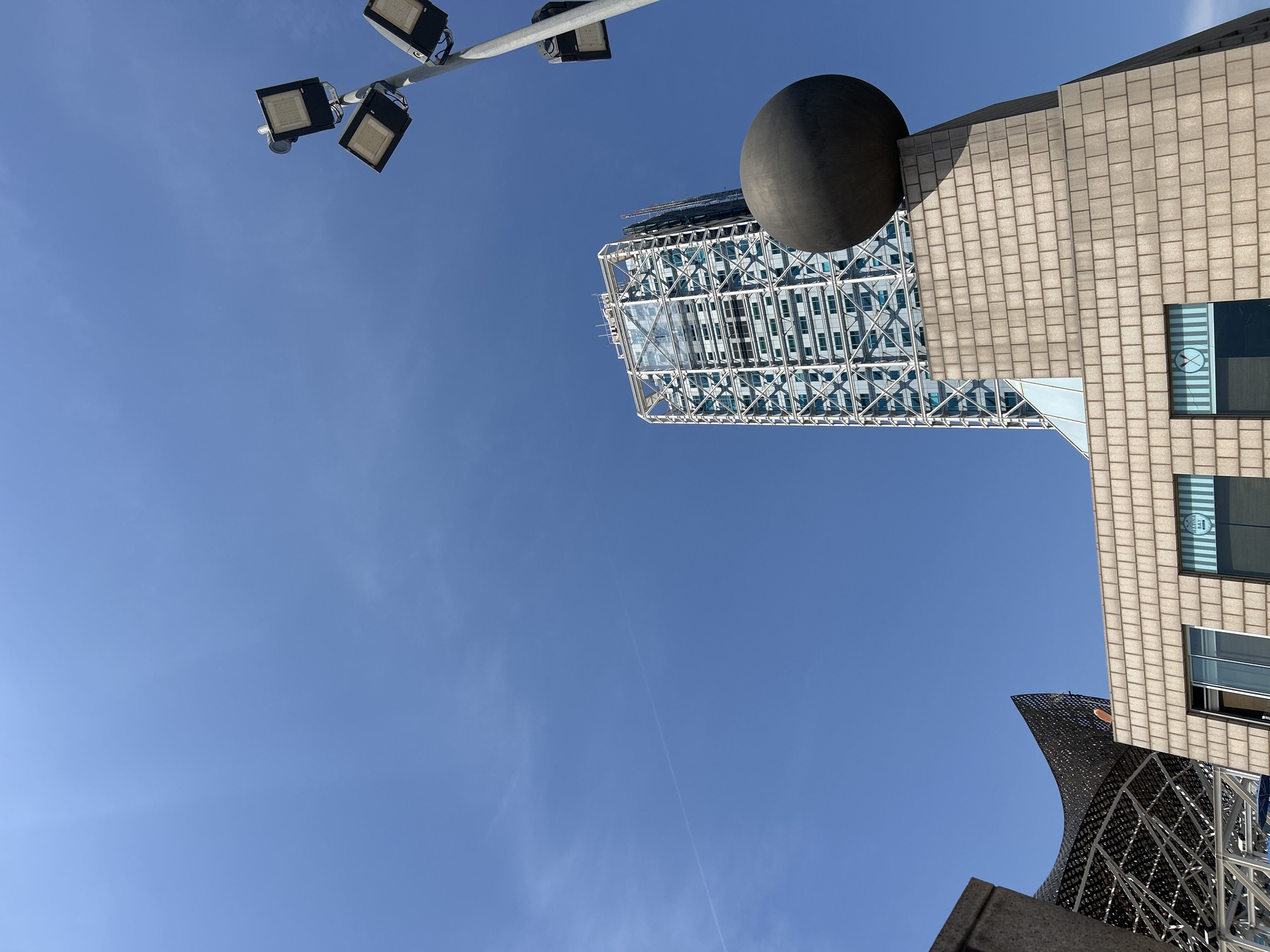 The image is a low-angle shot of a cityscape in Barcelona, Spain. The buildings are a mix of modern architecture, with one appearing to have a metallic cage-like exterior. A large dark sphere rests atop one of the structures. A light pole with multiple square floodlights is visible in the upper left corner. The sky is a clear, bright blue, with minimal cloud cover. The overall impression is urban, with a focus on architectural details.