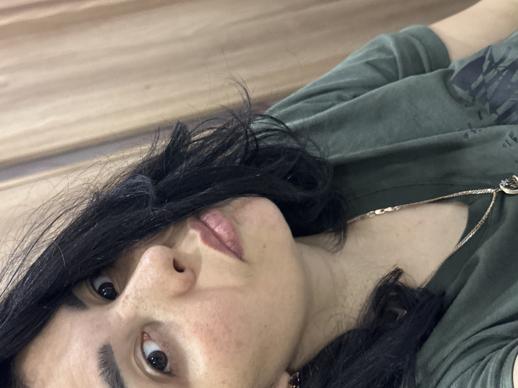 A close-up portrait of a person with dark hair. The person is wearing a green shirt and a gold necklace. The background appears to be a wood grain surface. The location and city are unknown.