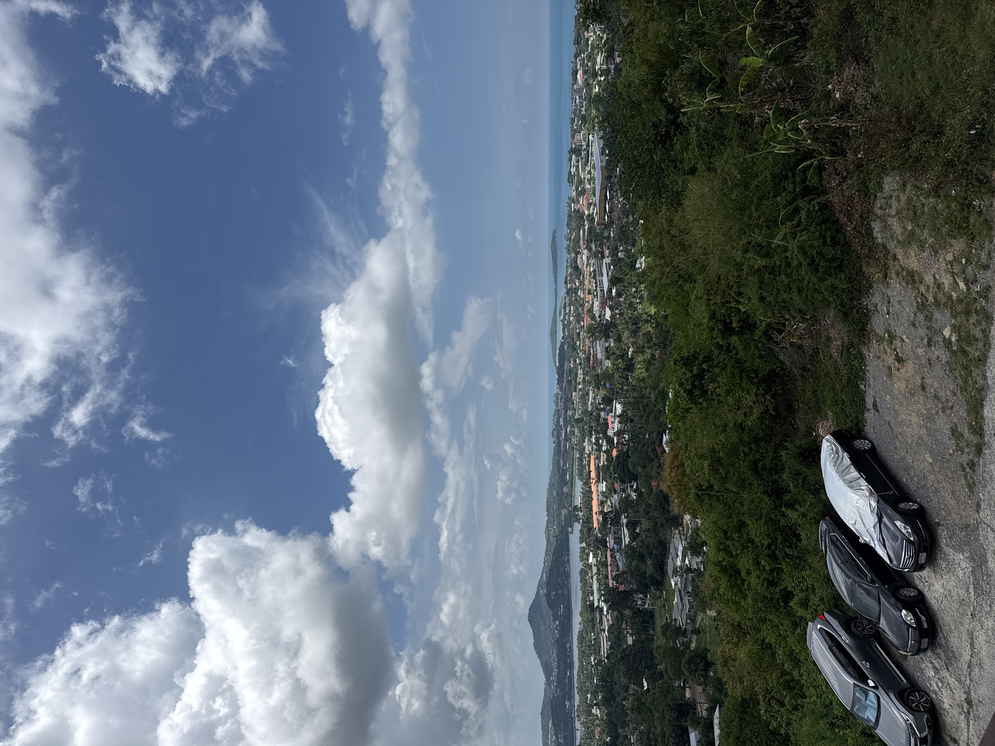 This is an outdoor, high-angle, full shot of a coastal city on a sunny day with scattered clouds. In the foreground, a gravel parking area holds three cars parked in a row from left to right: a dark gray SUV, a black sedan, and a black sedan covered with a reflective tarp. Behind the cars, dense green foliage covers a hillside. The midground shows a sprawling urban area with numerous buildings, some with orange tiled roofs, nestled amongst trees. A body of water, possibly a bay or inlet, separates the city from a distant green, mountainous island. The background is dominated by a bright blue sky filled with large, white cumulus clouds. The overall scene is peaceful and scenic.
