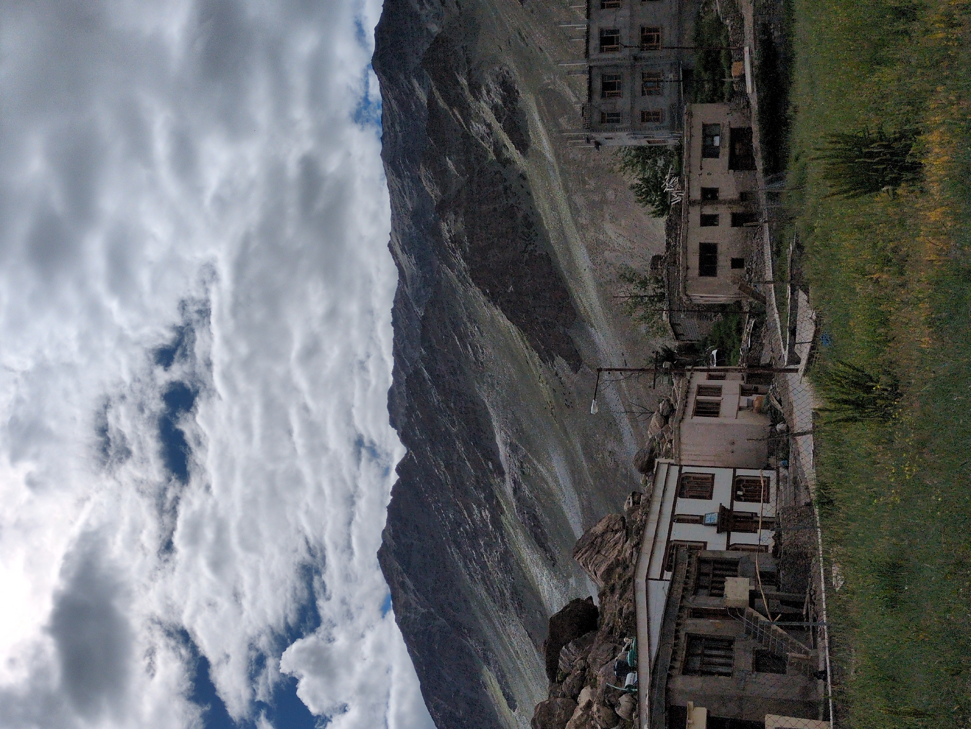 The image depicts a cloudy sky over a mountainous landscape with a village in the foreground. The village, located in Padum, India, features several buildings constructed with stone and concrete. Some of the structures appear to be residential, with windows and doors visible. One building, painted white with brown trim, has an entrance with a staircase. Another building, a multi-story concrete structure, has numerous windows. The surrounding environment is rugged, with rocky terrain and sparse vegetation, including tall grasses in the foreground. A street lamp is visible near one of the buildings.