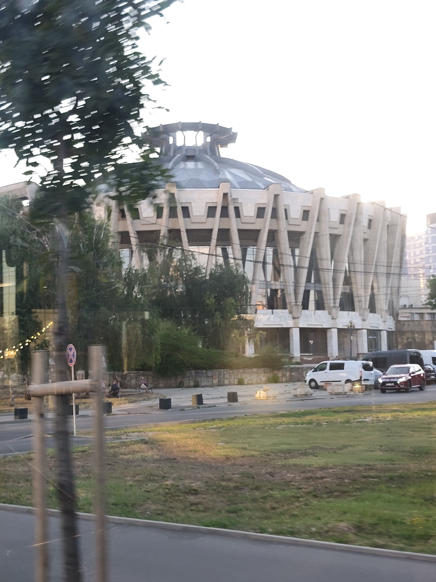 A large, circular, Brutalist-style building with a geodesic dome-like roof is visible from a street. The building's facade is characterized by dramatic, angular concrete supports. Trees and greenery partially obscure the lower part of the structure. Several vehicles, including a white van and a red SUV, are parked or driving on the street in front of the building. A pedestrian area with a few people sitting on benches is also present. The scene appears to be in an urban environment during daylight. This building is the Republic Palace (now the National Palace "Nicolae Sulac") in Chișinău, Moldova.