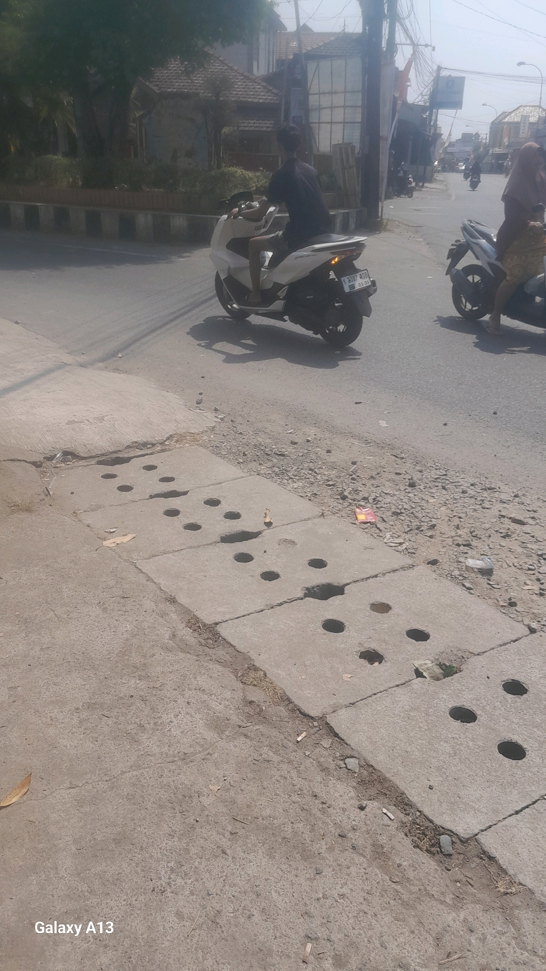The image depicts a street scene in Kirig, Indonesia. In the foreground, a concrete drainage system with circular openings is visible. The street is paved with asphalt, and a dirt and gravel shoulder runs alongside the road. In the middle ground, two individuals are riding motorcycles. The rider of a white scooter is wearing a black t-shirt and shorts, and the scooter has a license plate that reads "K 3387 AG." The rider of a darker motorcycle, partially obscured, is wearing a headscarf and a patterned skirt. In the background, buildings with tiled roofs and various structures line the street, suggesting a residential or commercial area. Trees and utility poles are also present. The sky is bright, indicating daylight.