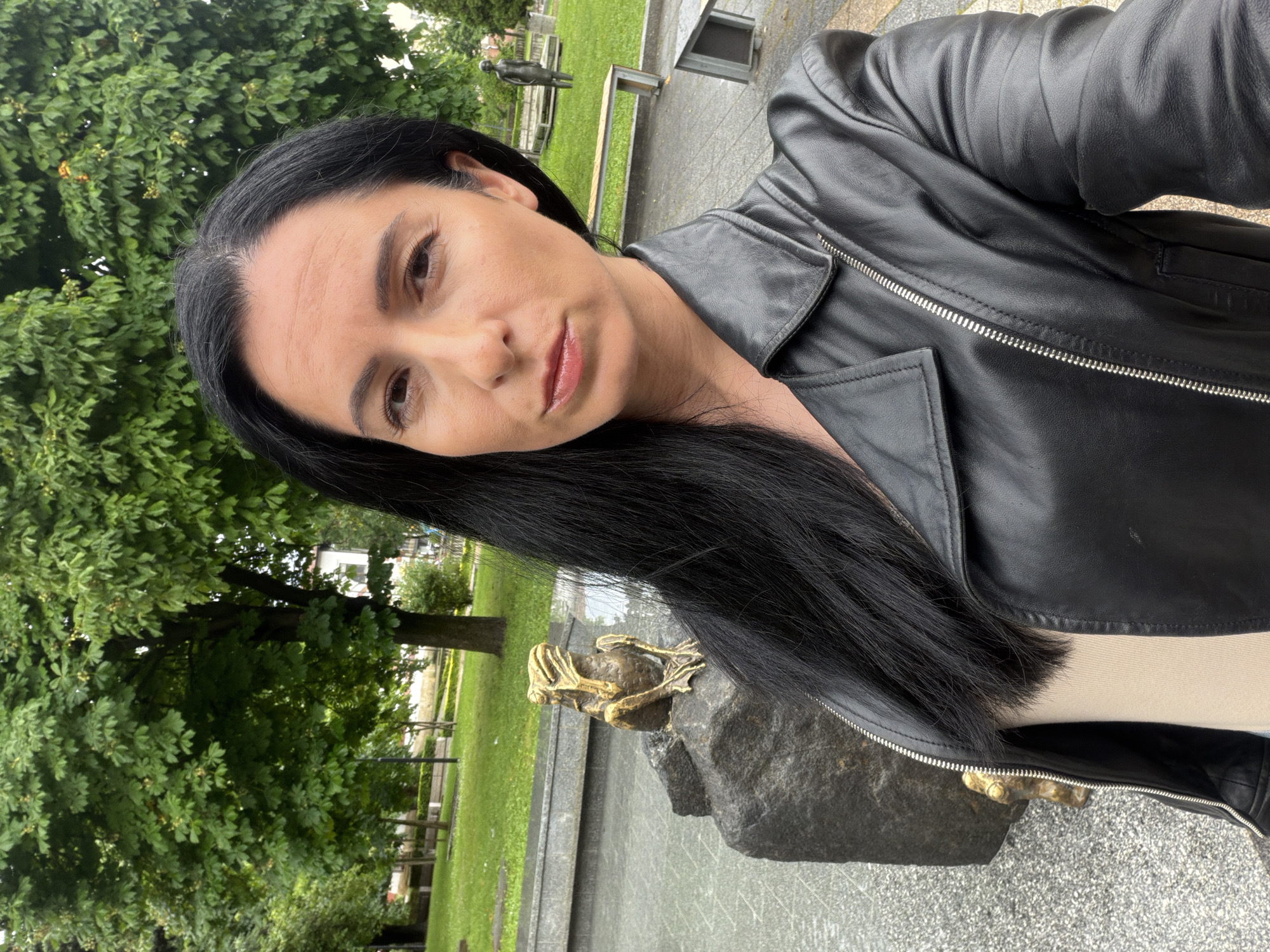 An outdoor, angled selfie-style portrait features a woman with long, straight dark hair and a black leather jacket, looking directly at the camera with a neutral expression. Her complexion is fair, and her eyebrows are dark and well-defined. She appears to be standing on a paved surface.
In the background, a distinctive bronze sculpture of a frog or toad-like creature, crouching on a dark, rough-hewn stone base, is visible behind the woman's left shoulder. This sculpture is a notable feature of the scene. The setting is a verdant park or public square, characterized by lush green trees with dense foliage, areas of green grass, and paved pathways. Further in the background, a smaller, recumbent human-like sculpture and a park bench can be discerned amongst the greenery.
The scene is lit by soft, natural daylight, suggesting either an overcast sky or the softer light of early morning or late afternoon, as there are no harsh shadows. The overall impression is one of a calm, pleasant outdoor environment. The location context provided indicates this scene is in Pleven, Bulgaria. No visible text is present in the image.