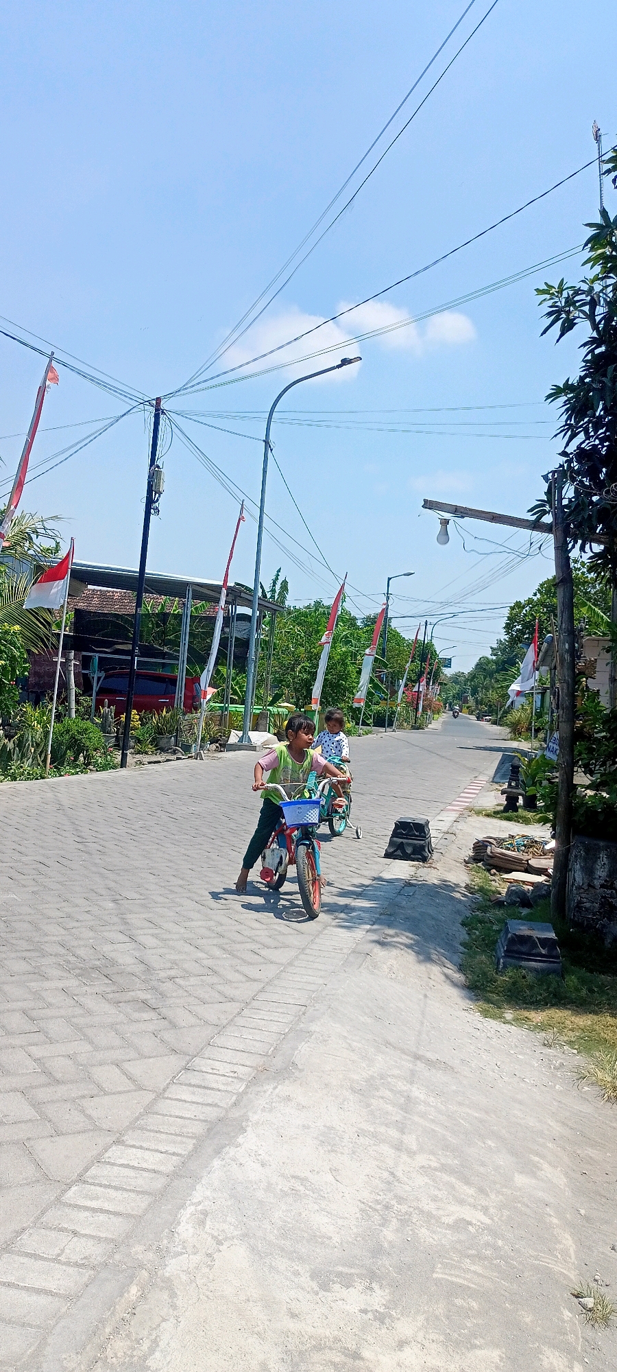 In Gempolklutuk, Indonesia, two young children are riding bicycles on a paved street lined with Indonesian flags. The child in the foreground, a girl wearing a green shirt and dark pants, is on a red and blue bicycle with a blue basket. She is looking forward and appears to be in motion. Behind her, another child, possibly a boy, is on a teal bicycle with training wheels. The street is made of interlocking gray pavers, and to the right, there is a dirt embankment with some debris. In the background, there are trees, houses, and electrical poles with numerous wires. The sky is clear and blue with a few wispy clouds.