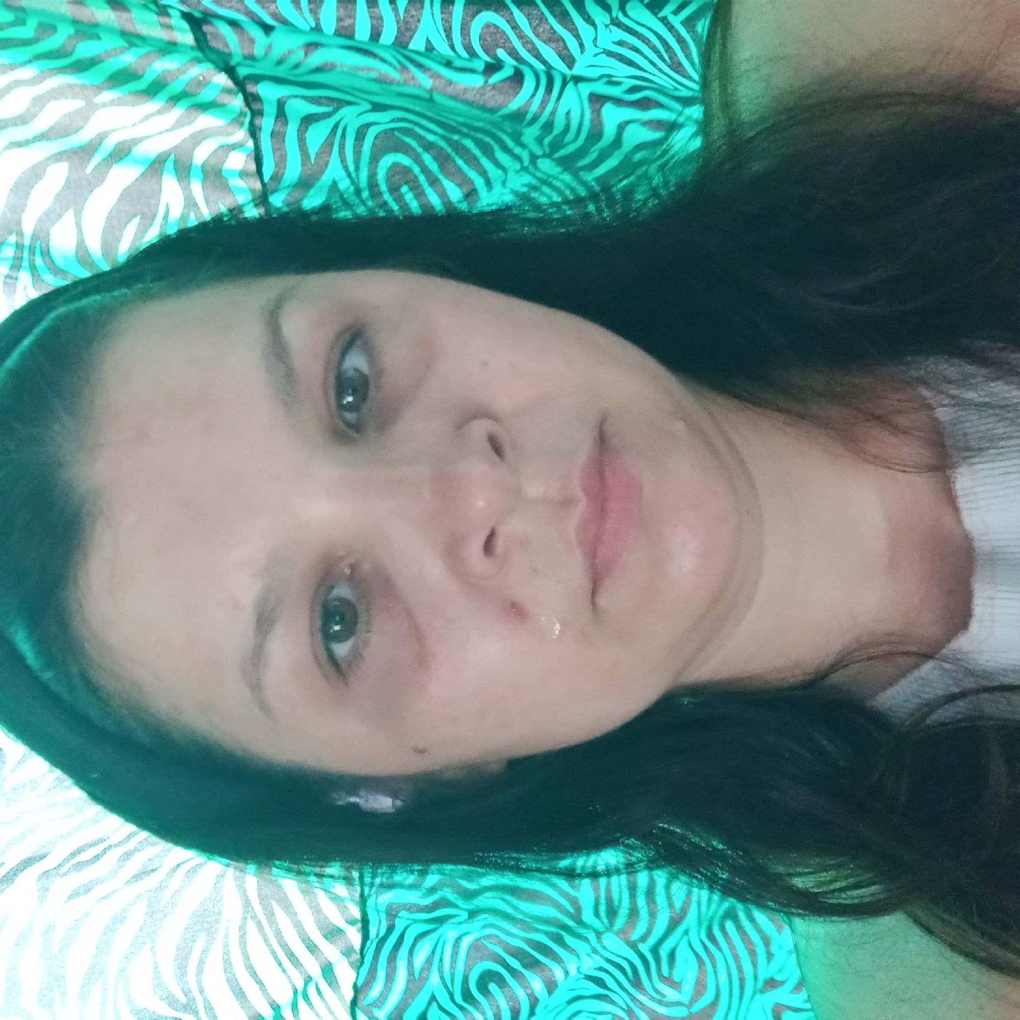 The image shows a medium close-up of a woman's face against a teal and black zebra-striped background. The woman has dark hair pulled back with a headband and appears to have a tear or wetness on her cheek and chin. There are dark circles under her eyes and a small blemish on her left cheek. Her gaze is directed forward. The lighting creates a green tint on the left side of the image.