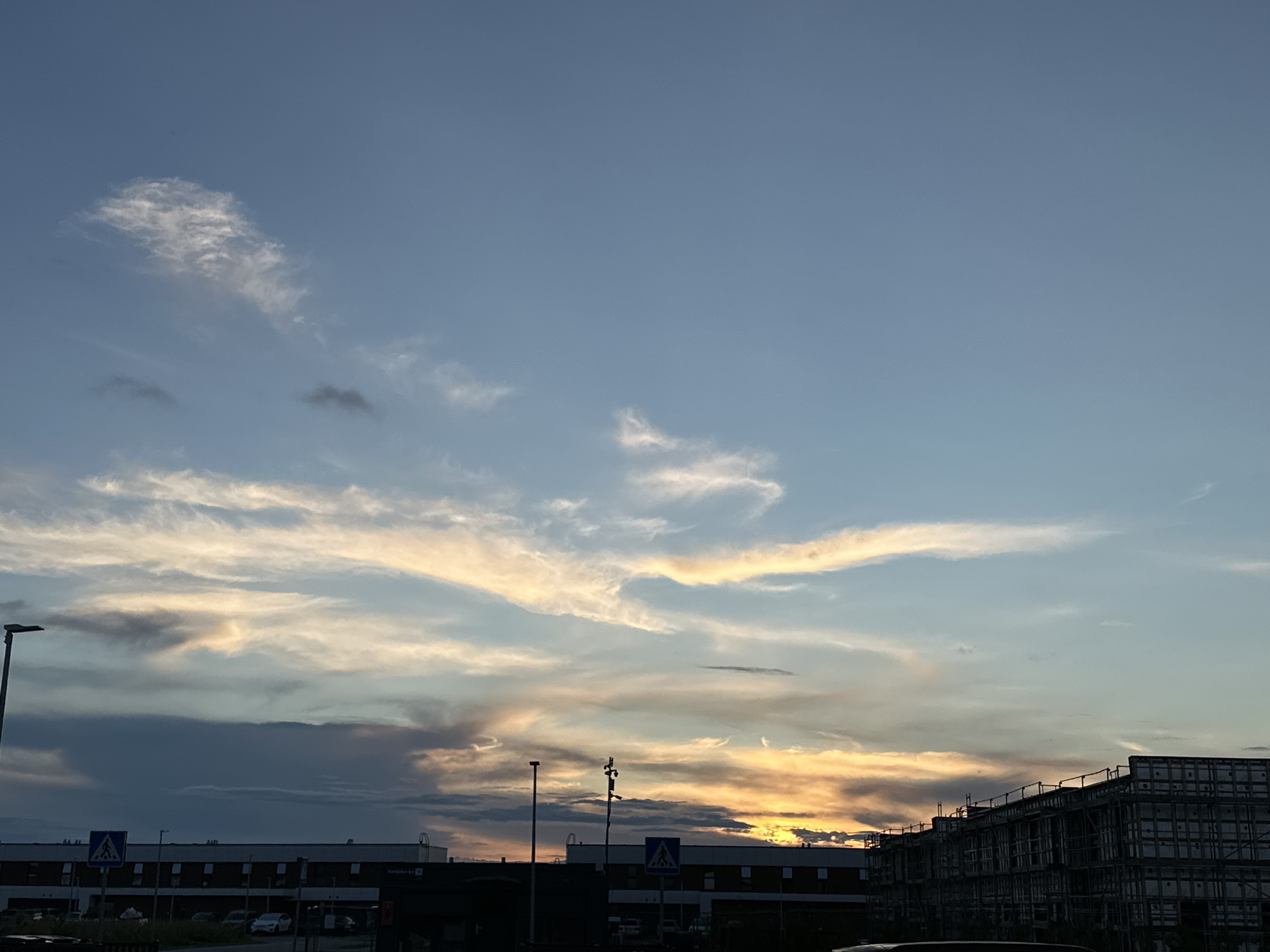 The image depicts a sky at dusk over a commercial or industrial area, likely in Tiskre, Estonia. The sky is a blend of blue and orange hues, with wispy clouds illuminated by the setting sun. Several buildings are visible, including a long, dark structure with many windows in the foreground and another building under construction to the right, characterized by scaffolding. A street lamp and a pedestrian crossing sign are also present. The scene appears to be a parking lot or street in front of these buildings. There are no people or animals clearly visible in the image.