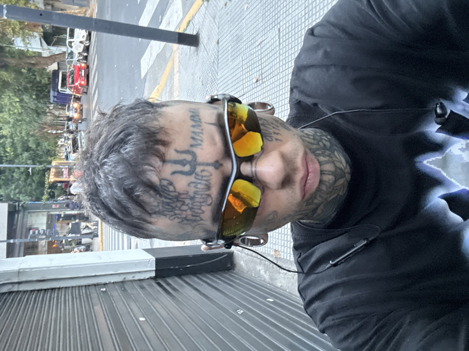 A person with extensive tattoos on their face and neck is captured at eye-level. The tattoos include intricate designs and lettering above the eyebrows. They are wearing sunglasses with reflective lenses. Large-gauge earrings are visible. They wear a black shirt with a lightning bolt graphic. The setting appears to be an urban street with a sidewalk featuring a gray textured surface. Cars, buildings, and trees are blurred in the background, suggesting a busy street scene.