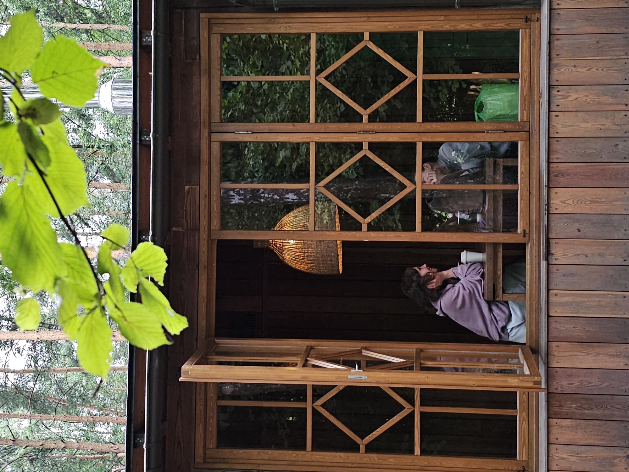 A close-up shot features a wooden cabin with large windows overlooking a forest. In the foreground, bright green leaves from a tree branch hang over the top of the cabin, obscuring the sky and a section of the roof. Through the open left pane of the large wooden-framed window, a young woman with brown hair is seated at a wooden table, resting her chin on her hand and looking to her right. She is wearing a light purple hoodie and light blue pants, and a white paper cup sits on the table in front of her. To her right, inside the cabin, a woven lampshade hangs from the ceiling. In the right section of the window, a person is visible, but their features are obscured by shadow and a large ceramic vase on the table. A bright green plastic bag is placed on the floor behind them. The exterior of the cabin is clad in vertical wooden planks, with the window frames made of polished wood. The forest visible through the windows consists of tall, slender pine trees and dense green foliage. The scene appears to be set in Nemchinovo, Russia, a village known for its natural surroundings.