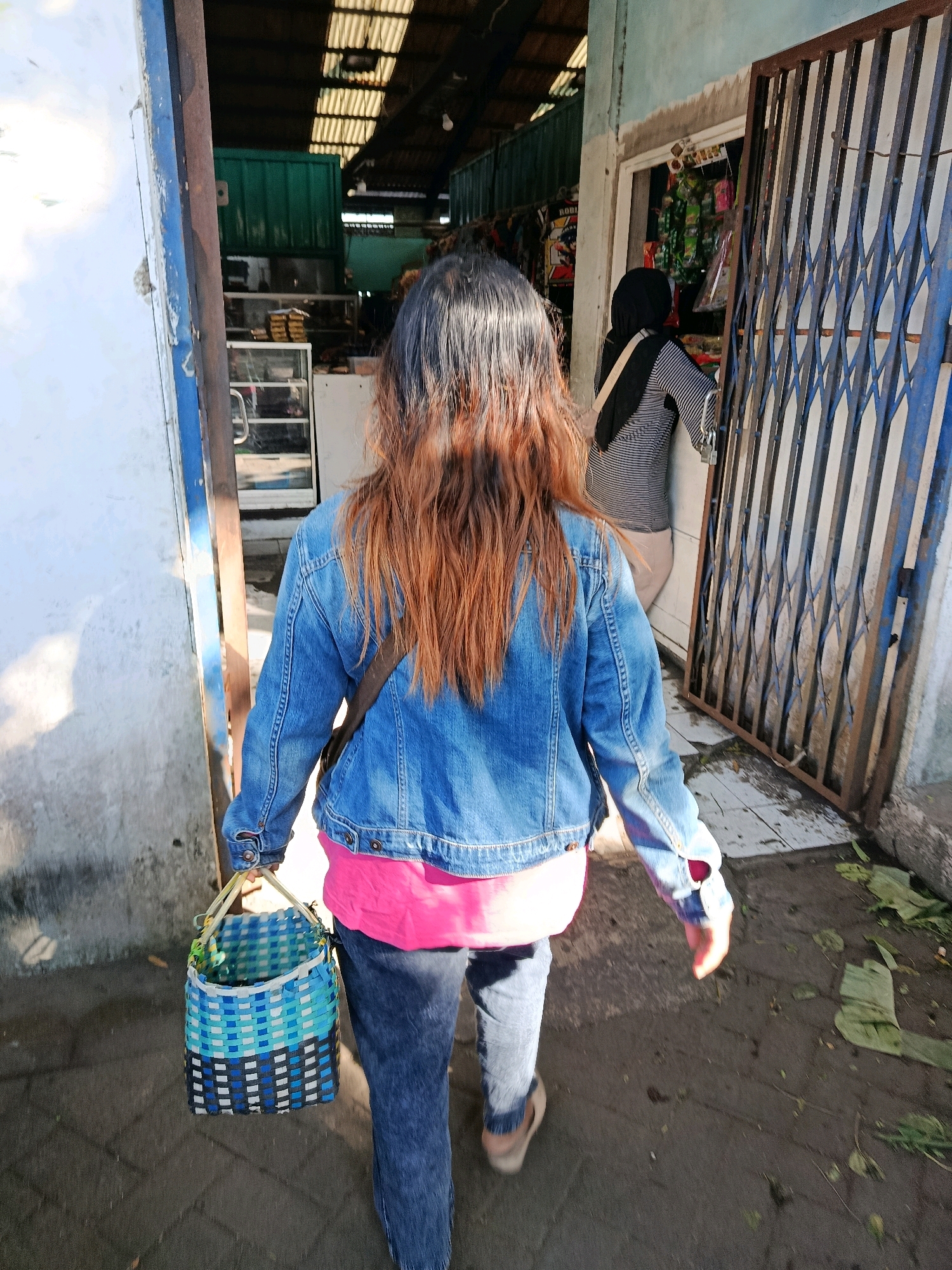 A person walks away from the camera, holding a woven basket. They are wearing a denim jacket over a pink shirt and blue jeans. The basket is primarily blue and white with black accents. The person is walking towards an open doorway with a metal gate on the right. Inside the doorway, another person wearing a black hijab and a striped shirt is visible, appearing to interact with items on display. The scene appears to be a market or shop, with stalls and goods visible in the background. The ground is paved, and there are some scattered leaves and debris. The overall lighting suggests it is daytime, with sunlight casting shadows. The location is identified as Bululawang, Indonesia.