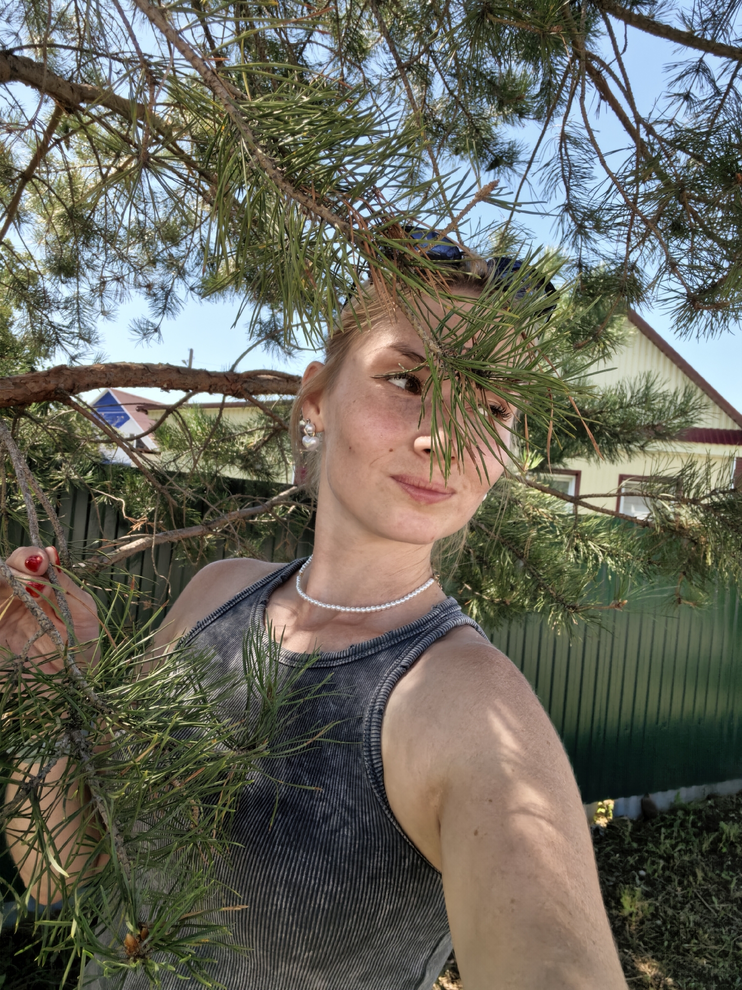 A young woman is photographed outdoors, partially obscured by pine branches. She has blonde hair pulled back, fair skin with freckles, and is wearing a dark grey ribbed tank top, a pearl necklace, and earrings. Her right hand, with red-painted fingernails, is touching a pine branch. In the background, a light-colored house with a red roof and a green fence are visible. The sky is clear and blue. The image appears to be taken on a sunny day.