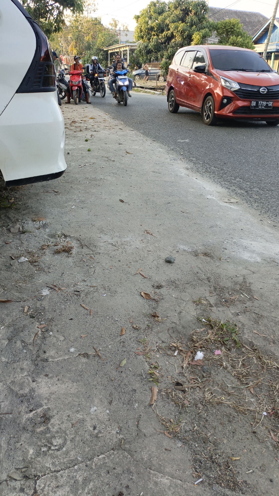 The image depicts a street scene in Gusunge, Indonesia. In the foreground, a white car's rear is partially visible on the left. The ground is a dusty, uneven surface with scattered dry leaves and small stones. To the right, a paved road is visible with an orange car driving on it. Further down the road, several people on motorcycles are present. One red motorcycle has two riders, and a blue motorcycle has two riders, including a child passenger. In the background, trees and buildings line the street. The lighting suggests it is daytime, possibly late afternoon due to the warm tones.