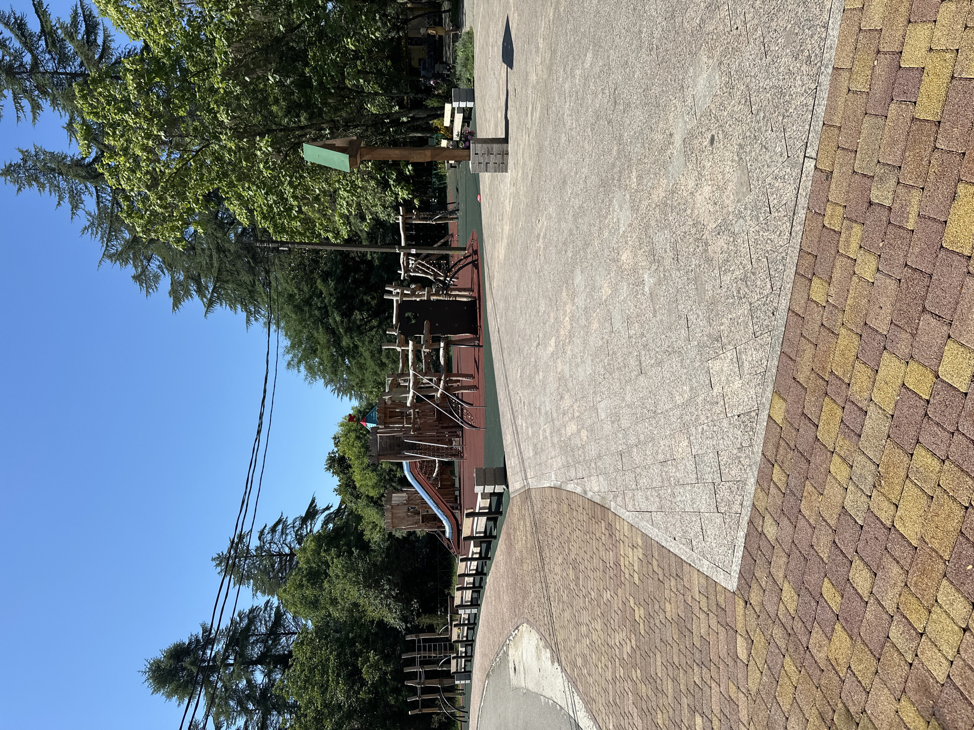 This image depicts a modern, well-maintained children's playground located in Sochi, Russia. The playground features a large wooden climbing structure with a blue slide, various ladders, and net climbs. Surrounding the play area are benches for seating, all set against a backdrop of lush green trees and a clear blue sky. The ground is paved with interlocking bricks in a pattern of red and yellow, transitioning to a lighter, speckled stone-like material in other areas. In the foreground, the paving stones are clearly visible, forming a path that curves around the playground. The scene is bathed in bright sunlight, casting shadows on the ground, suggesting a warm, pleasant day. There are no people visibly interacting with the playground equipment in this particular shot, though a person can be seen in the distance, standing near some benches. The overall impression is one of a peaceful, recreational space designed for children's enjoyment in a natural setting.