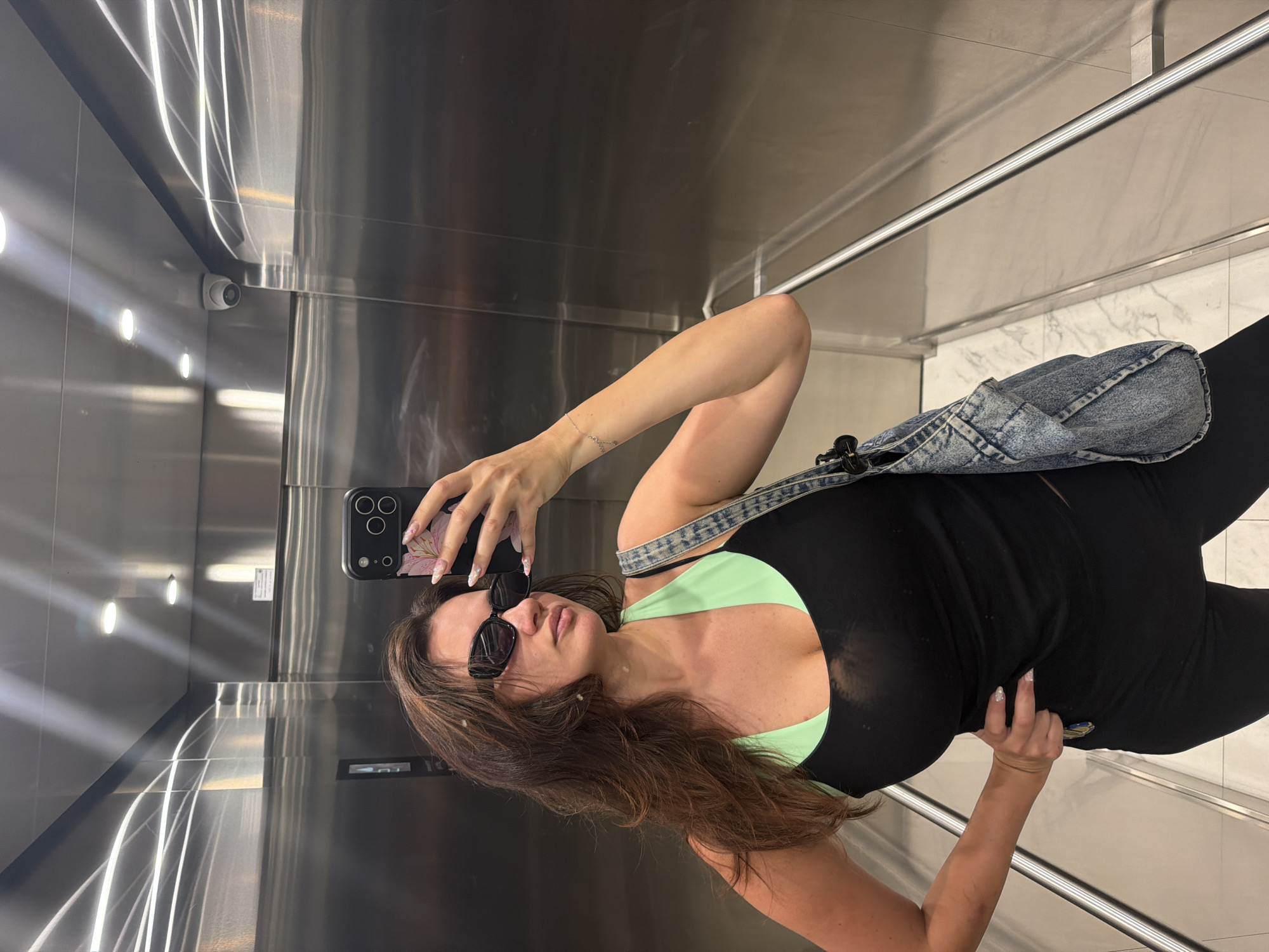 A woman is taking a mirror selfie in an elevator. She is wearing sunglasses, a black top with a green halter neckline, and a denim bag is slung over her shoulder. Her long, brown hair is down, and she is holding a smartphone in her left hand. The elevator walls are made of reflective stainless steel, with lights creating streaks of light on the surface. A security camera is visible on the wall. The elevator also features a chrome handrail and marble-patterned flooring. The overall mood appears casual and everyday. There is no visible text in the image. The setting suggests an indoor urban environment, likely in Belgrade, Serbia.