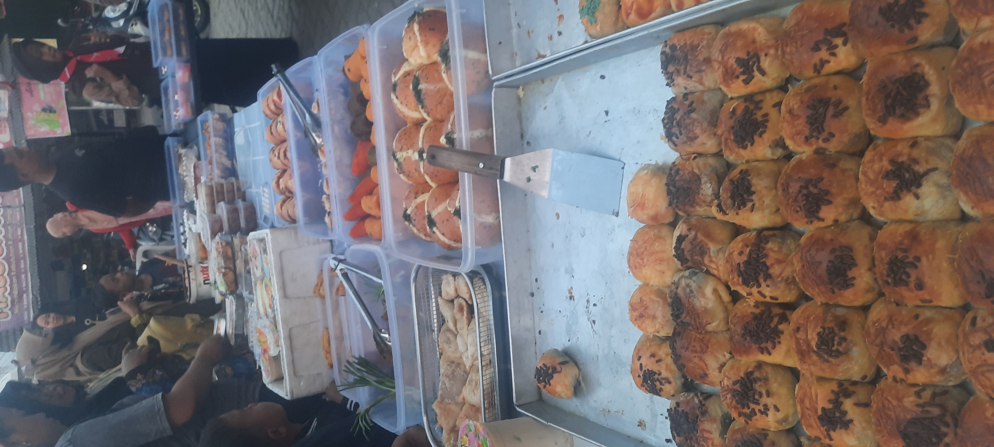 An outdoor market scene in Selopuro, Indonesia, features a food stall laden with various baked goods and snacks. Several individuals, including women in hijabs and children, are visible browsing or working at the stall. The foreground displays trays of small pastries, some topped with chocolate sprinkles and others with grated cheese. Behind these, rows of larger, round baked goods are arranged in plastic containers. Further back, more containers hold items like fried snacks, green onions, and what appear to be small peppers or carrots. A metal spatula rests on one of the trays. In the background, more people are present, and market stalls with banners suggest a bustling commercial environment. The lighting indicates it is likely daytime.