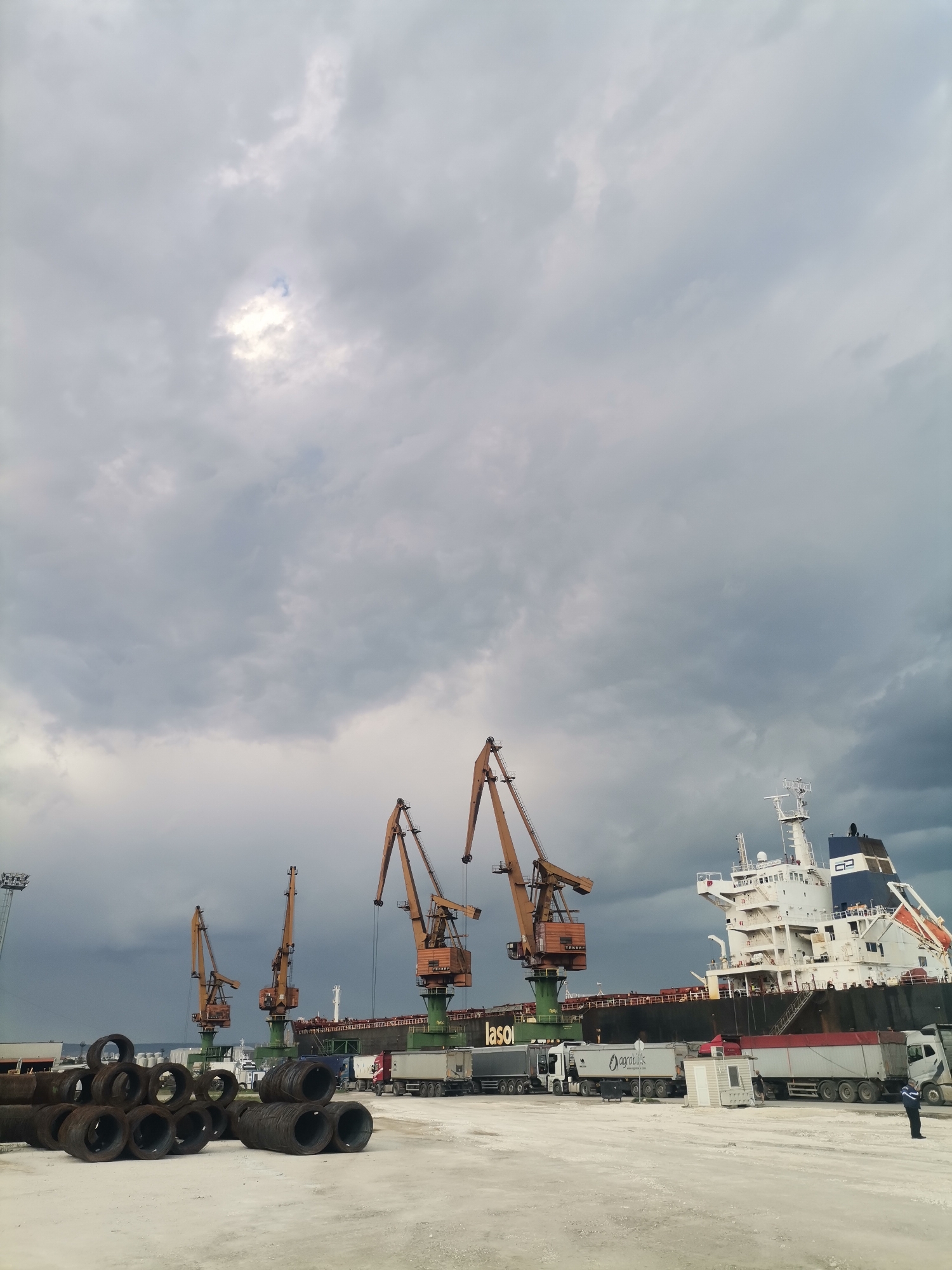 The image captures a scene at the port of Ezerovo, Bulgaria. Four large, yellow-orange cranes stand along the dock, positioned near a massive cargo ship with a white and blue superstructure. Semi-trailer trucks are parked alongside the ship, suggesting ongoing loading or unloading operations. In the foreground, stacks of coiled metal products, likely steel, are visible. A lone individual is standing near the side of the ship; their uniform is visible. The sky above is overcast with heavy, dark clouds, hinting at impending or recent rain. The ground is a light color, appearing to be concrete or a similar material typical of industrial port areas.
