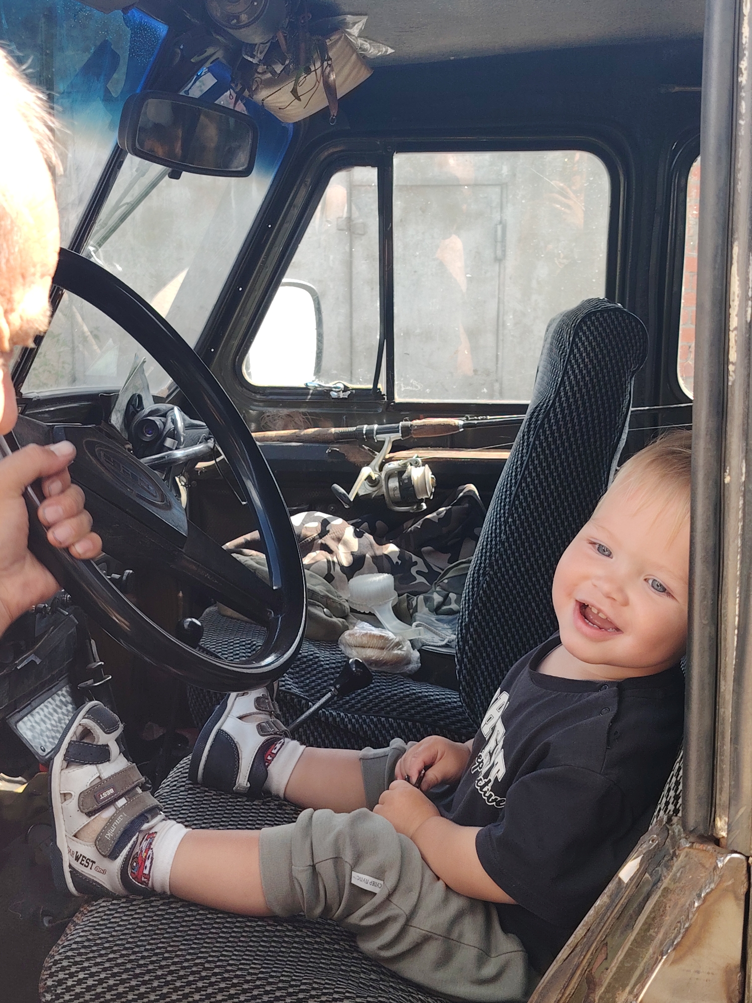The image shows a young child sitting in the driver's seat of a vehicle, possibly a truck, in what appears to be an outdoor setting. The child, who is wearing a black t-shirt, grey pants, and sandals, is smiling broadly and looking towards the camera. The child's feet are resting on the seat. The steering wheel is in the foreground, and the interior of the vehicle is visible, including a seat and some fishing rods. The background is out of focus but appears to be an outdoor area. The lighting suggests it is daytime. The scene is likely in Chaikovsky, Russia, as indicated by the provided location.