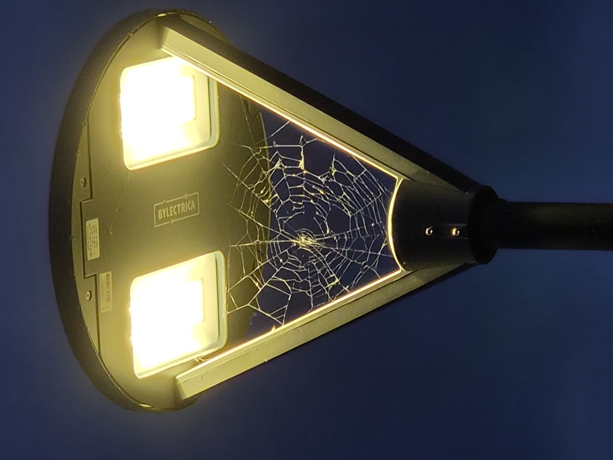 The image shows a street lamp at night. The lamp has two bright yellow lights and a cracked, web-like structure is visible in the center, possibly a spiderweb. The background is a dark blue sky. Text "BYSELECTRICA" is visible on the lamp housing. No people or animals are visible in the image. This street lamp is located in Zhodzina, Belarus.