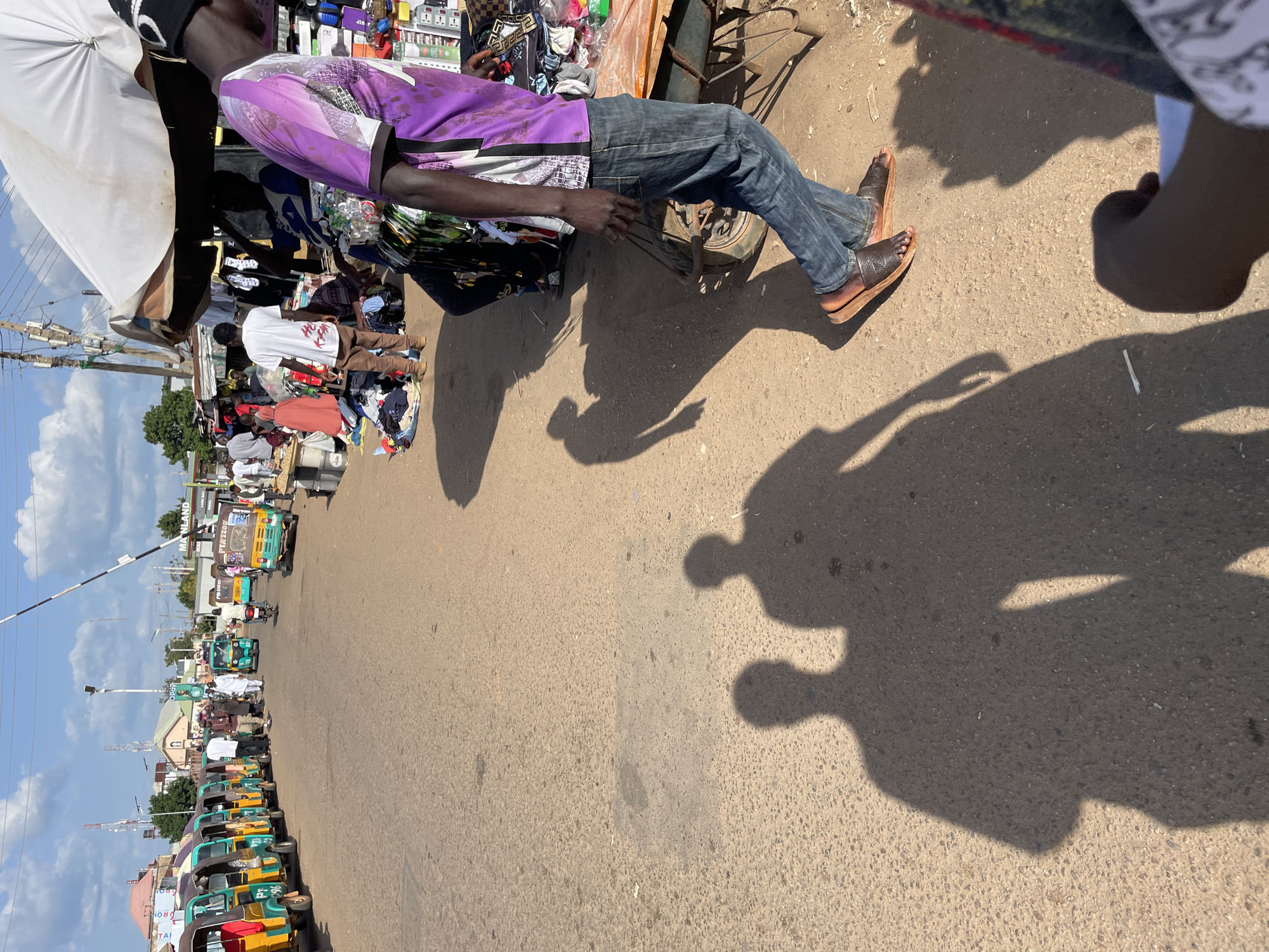 The image depicts a street scene in Bukuru, Nigeria, likely a market area. A line of colorful auto-rickshaws (keke-napeps) is parked on the side of a paved road. Several people are walking and standing in the vicinity of the vehicles and market stalls.
In the foreground, the shadows of two individuals are cast on the road, indicating the direction of the sun. A man wearing a purple patterned shirt and blue jeans is walking towards the right of the frame, his shadow also visible. He is carrying a wheelbarrow with a handle. Behind him, market stalls display various goods, including clothing and other merchandise.
Further down the street, more people are visible, some interacting with vendors. A white tent or umbrella provides shade over one of the stalls. In the background, buildings and telecommunication towers can be seen against a clear sky with scattered clouds. The overall impression is that of a busy, open-air market on a sunny day.