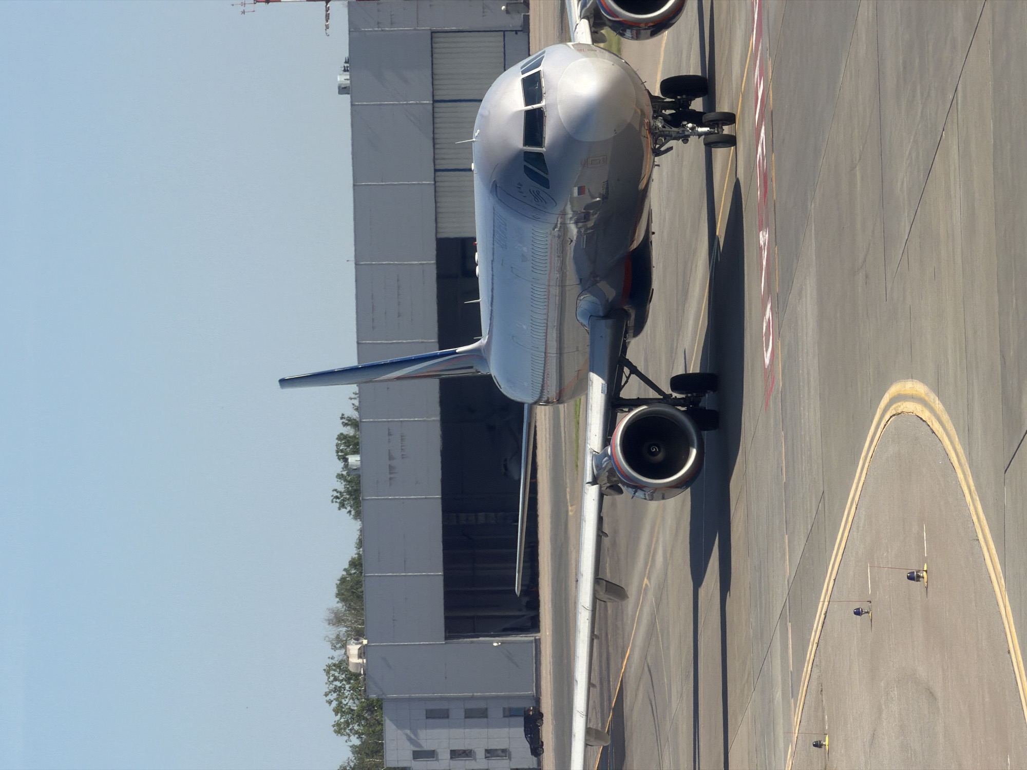 An Aeroflot Airbus A320 aircraft is on the taxiway at Sheremetyevo International Airport (SVO) in Khimki, Russia. The aircraft is silver with blue and red accents on the tail. It is facing right, with its nose pointed towards the right side of the frame. The wings are extended, and the engines are visible under the wings. The aircraft is on a paved taxiway with yellow lines marking the edges and red markings on the ground in front of the aircraft. In the background, a large hangar building is visible, with its doors open. There are trees and some other buildings behind the hangar. A dark-colored car is parked in front of the hangar. The sky is clear and blue.