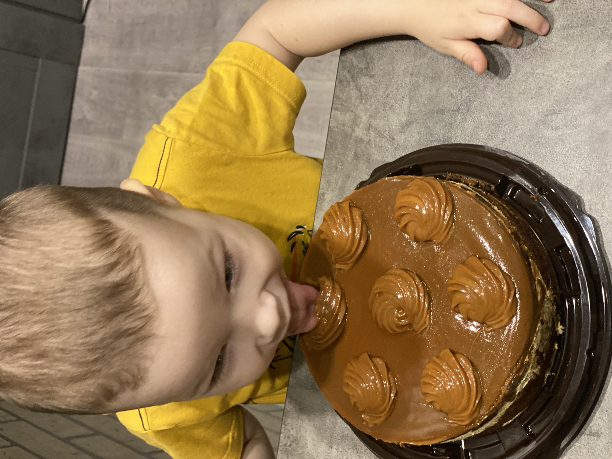 A young child in a yellow t-shirt is licking the icing off a caramel cake, which is sitting in its brown plastic container on a gray surface. The child's hand is resting on the surface next to the cake. The location is not specified in the image and cannot be determined.