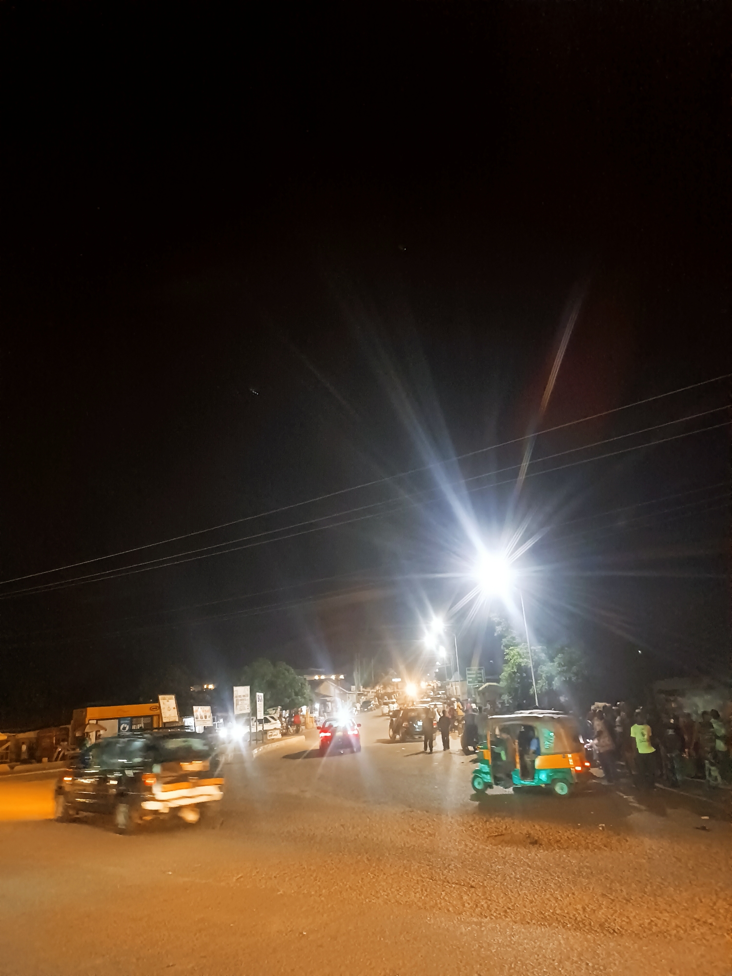 The image depicts a street scene at night in Bukuru, Nigeria. The scene is illuminated by bright streetlights, which create a glare. In the foreground, a car is traveling left to right. To the right of the car is a green and yellow auto rickshaw or "keke." A group of people is standing on the right side of the road. In the background, there are more vehicles, signs, and buildings. The sky is dark and clear with a few stars visible. Power lines crisscross the upper part of the frame.