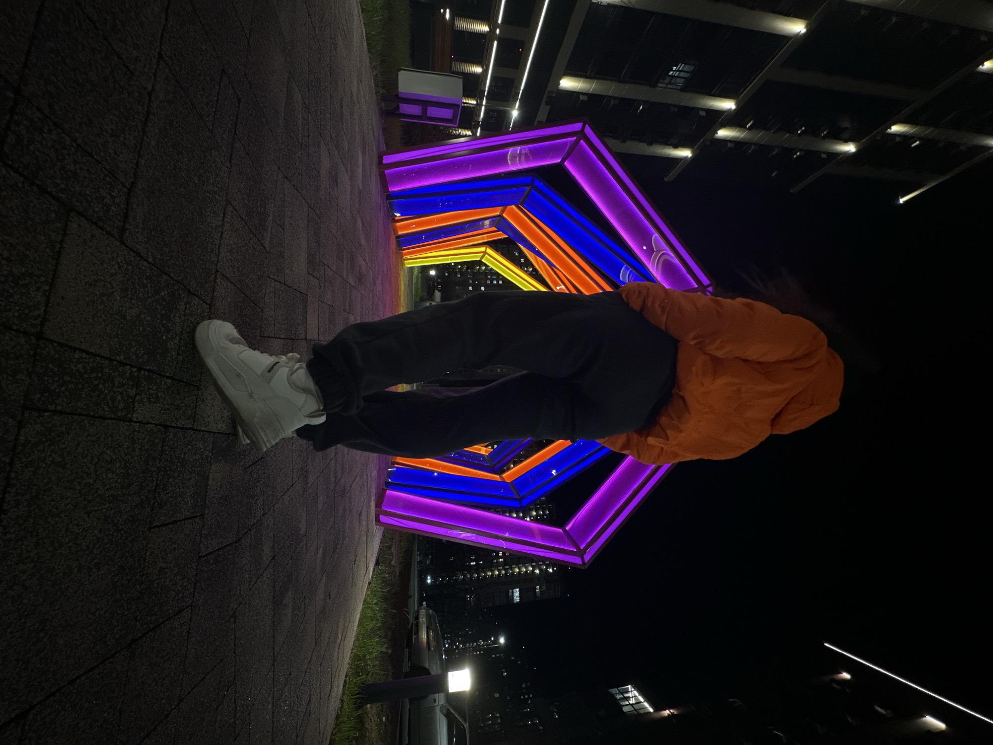 Here is a detailed description of the image:
**Scene Overview:**
The photograph, taken at night, depicts a person standing under a colorful, illuminated tunnel-like structure. The setting appears to be urban, with buildings visible in the background.
**People and Actions:**
A person is the central subject. They are wearing white sneakers, dark pants, and an orange jacket. The person is standing, seemingly posing, within the tunnel.
**Objects and Environment:**
* **Illuminated Tunnel:** The primary object of interest is the tunnel structure. It is comprised of a series of repeating geometric shapes that appear to be hexagonal or pentagonal. These shapes are lit up with vibrant colors, including purple, blue, orange, and yellow, creating a dynamic and eye-catching visual effect.
* **Urban Landscape:** The backdrop reveals an urban environment, with buildings of varying heights and lighting conditions.
* **Pavement:** The person is standing on a paved surface, likely a sidewalk or pedestrian walkway.
**Location and City:**
The image is taken in Rostov-on-Don, Russia.
