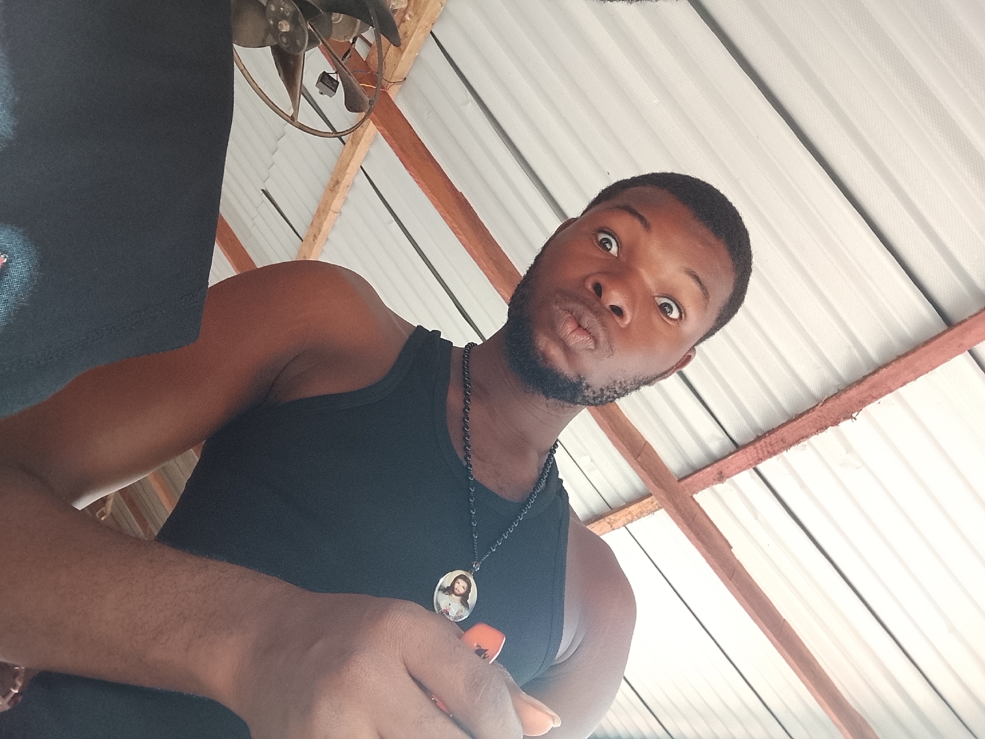 A low-angle selfie captures a young man with dark skin, a beard, and a mustache, making a humorous expression with wide-open eyes and pursed lips, looking directly at the camera. He is wearing a black tank top. Around his neck, he sports a black beaded necklace with a silver oval pendant. The pendant features an image, likely of Jesus Christ, depicted with long hair, a halo, and a red garment. His right hand, positioned in the foreground, holds an object that appears to be a phone with an orange case. The background consists of a bright, white corrugated metal ceiling supported by wooden beams. An old, industrial-style ceiling fan is partially visible in the upper left corner of the frame. The bright, even lighting suggests the photo was taken during the daytime in what appears to be a covered outdoor space or a simple indoor structure. There is no visible text in the image.