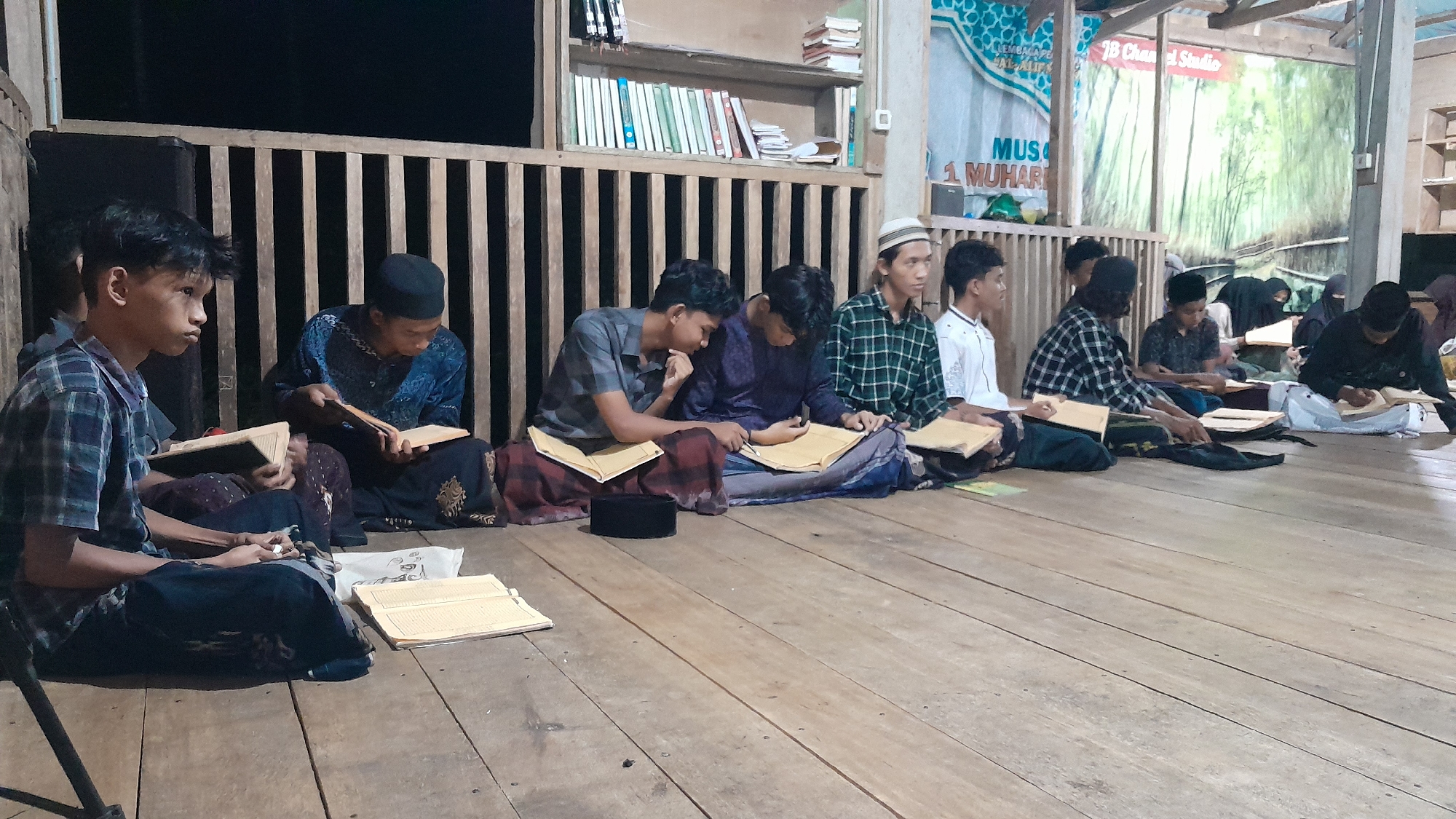 A group of young males, dressed in traditional attire, are sitting on a wooden floor, engaged in reading. They are positioned in a line, facing forward, with books open in front of them. Some wear skullcaps or turbans. The scene appears to be indoors, possibly in a mosque or madrasa, given the presence of bookshelves filled with religious texts in the background. The lighting is dim, suggesting it might be evening or nighttime. The wooden planks of the floor and the rustic wooden structure behind the individuals provide a sense of place. The overall atmosphere seems peaceful and studious, with the individuals focused on their reading. A banner with text is visible in the background, partially obscured, but some words like "MUS" and "1 MUHAR" are discernible, along with a logo that includes "JB Chan el Studio". The location is Lhokjok, Indonesia.