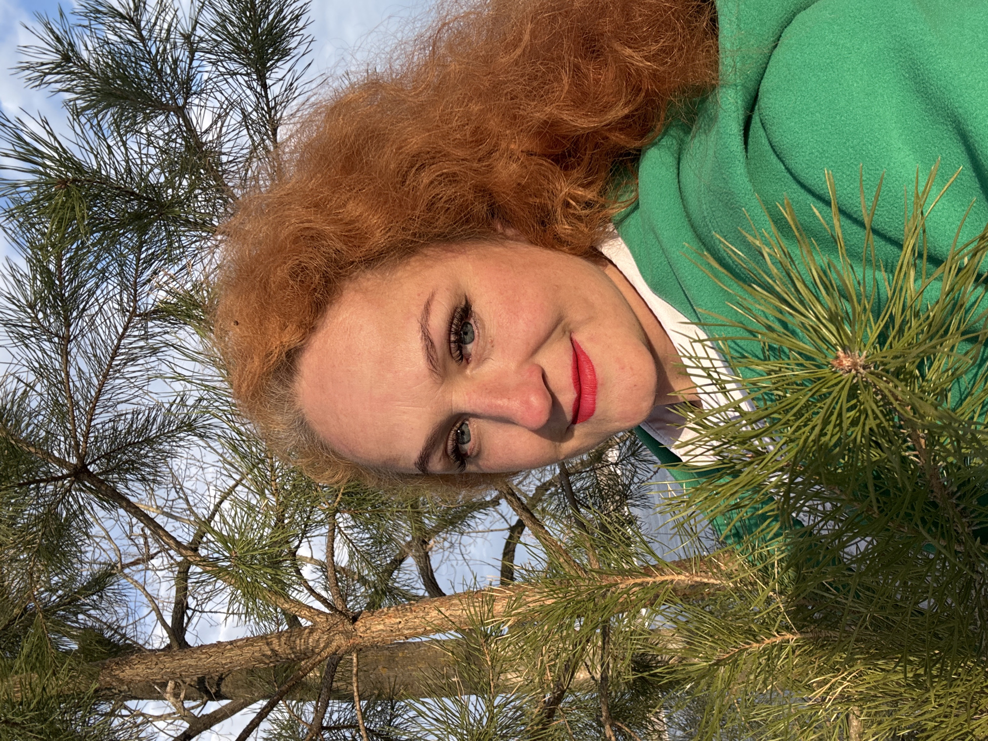 A woman with red curly hair, wearing a green jacket and white shirt, is positioned in front of a pine tree. The camera angle is low, looking up at the woman, who is smiling. The background consists of pine branches and a partly cloudy sky. The photograph appears to be taken outdoors, likely in a park or forested area in Noviy, Russia, based on the location provided.