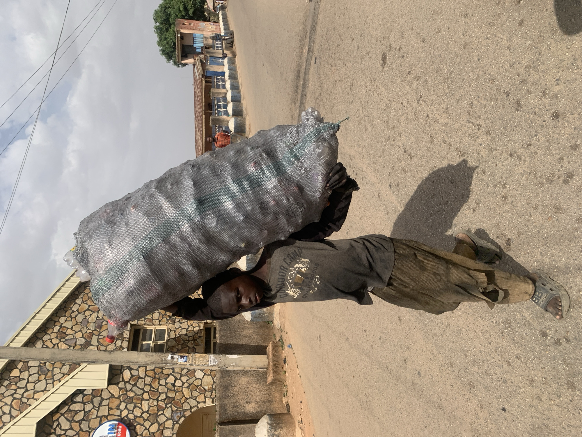 A young person, likely a boy, is depicted in a daytime street scene in Jos, Nigeria, carrying an extremely large, grey mesh bag filled with clear plastic bottles. The bulky load is balanced on his head and shoulders as he walks on an asphalt road. He wears a dark, faded t-shirt with the visible text "OUTDOOR CAMP" and "QUALITY GOODS." His dark trousers are worn, with a tear visible on the right leg, and he wears dark sandals, one of which clearly shows the brand "BALENCIAGA" on its strap. His facial expression appears focused, suggesting the effort involved in carrying the heavy bag.
The background features an urban environment under an overcast or partly cloudy sky, with power lines visible in the upper left. To the left, a building with a distinctive facade of multi-toned stone (brown, grey, and beige) and a light-colored roofline is visible. A partially obscured circular blue and white logo is on this building. A concrete utility pole covered with numerous posters or flyers stands near the stone-clad building. Further along the street on the right, another building with a reddish-brown wall and blue-shuttered windows can be seen. The road surface is dry asphalt, and faint shadows suggest it is daytime.