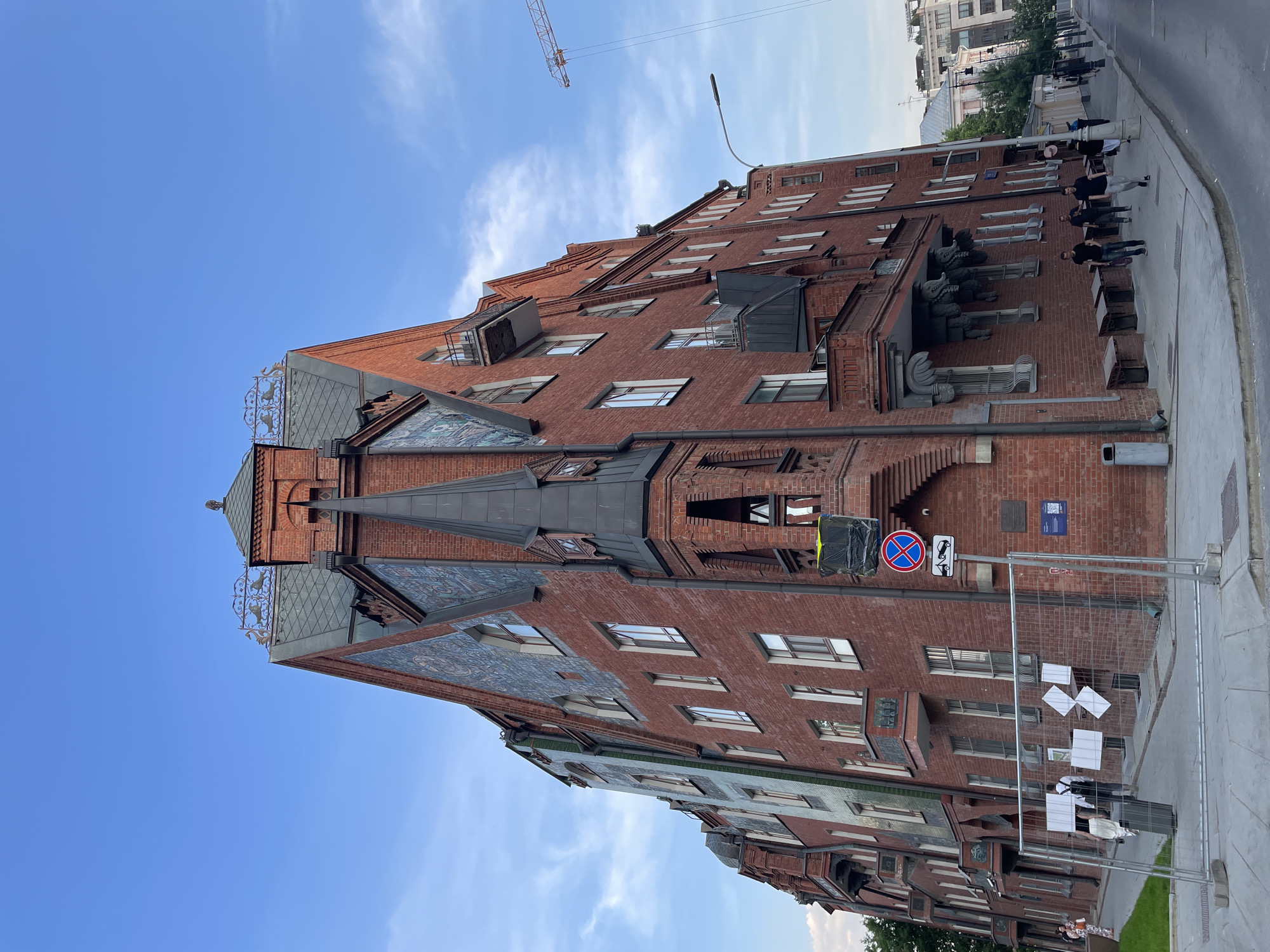The image captures a red brick building under a blue sky with some clouds. The building has multiple floors and an ornate architectural design, including tiled patterns on parts of the roof and facade. On the building's facade, there are statues of figures along the sidewalk level. A construction crane is visible in the background, slightly blurred. Some people are walking along the sidewalk. A street sign is posted near the building's entrance. A barrier is set up on the sidewalk next to a set of postings on paper. The location is in a city that is identified as Unknown, Unknown.