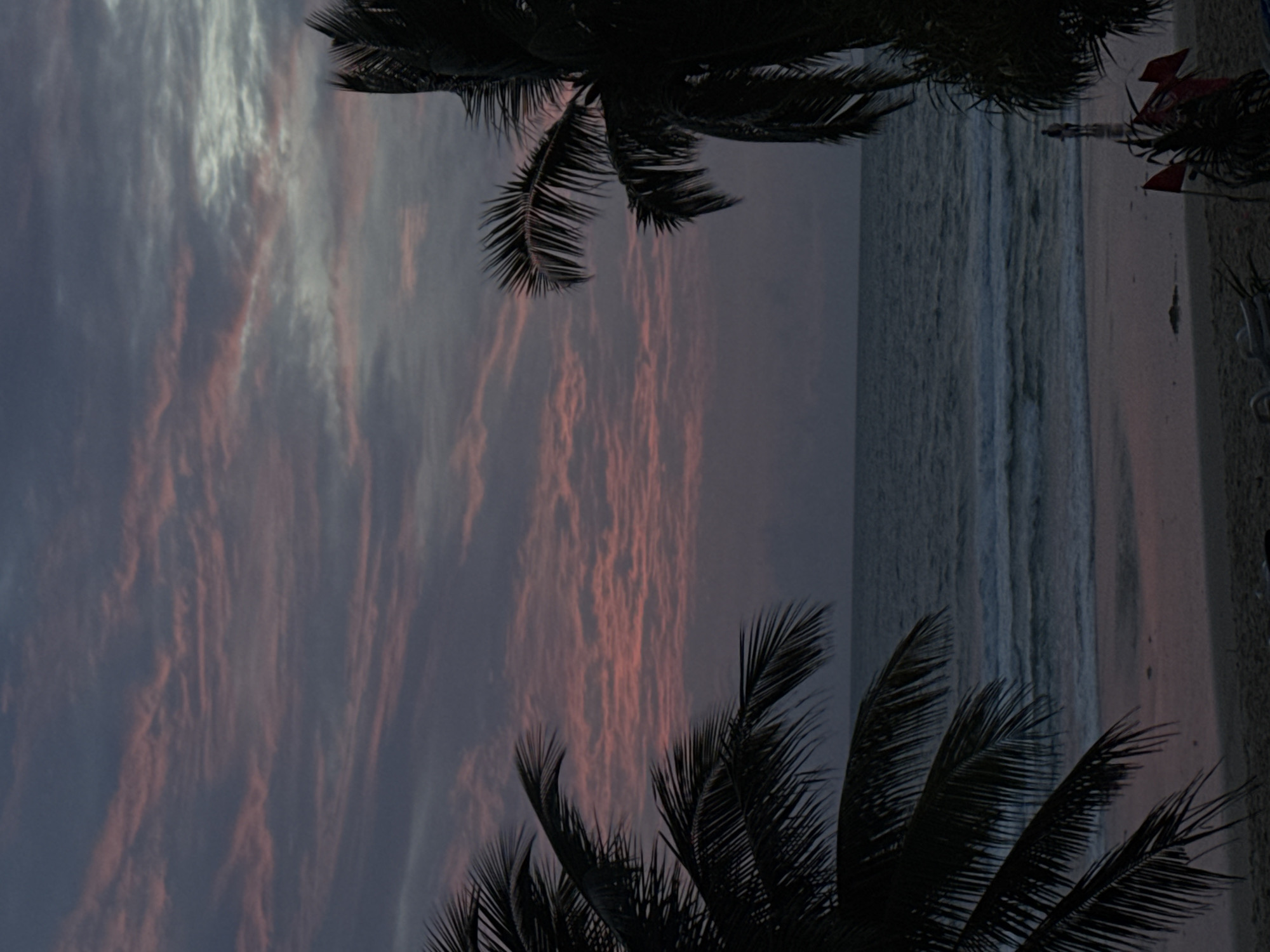 The image depicts a serene sunset scene at Karon Beach, Thailand. The sky is painted with hues of pink and purple, with wispy clouds stretching across the horizon. Palm trees frame the view from both sides, their silhouettes stark against the colorful sky. The calm ocean laps gently onto the sandy shore, where a few distant figures are visible walking near the water's edge. Red flags are planted in the sand, adding a touch of color to the otherwise muted tones of the beach. The overall atmosphere is tranquil and picturesque, capturing the beauty of a tropical evening.