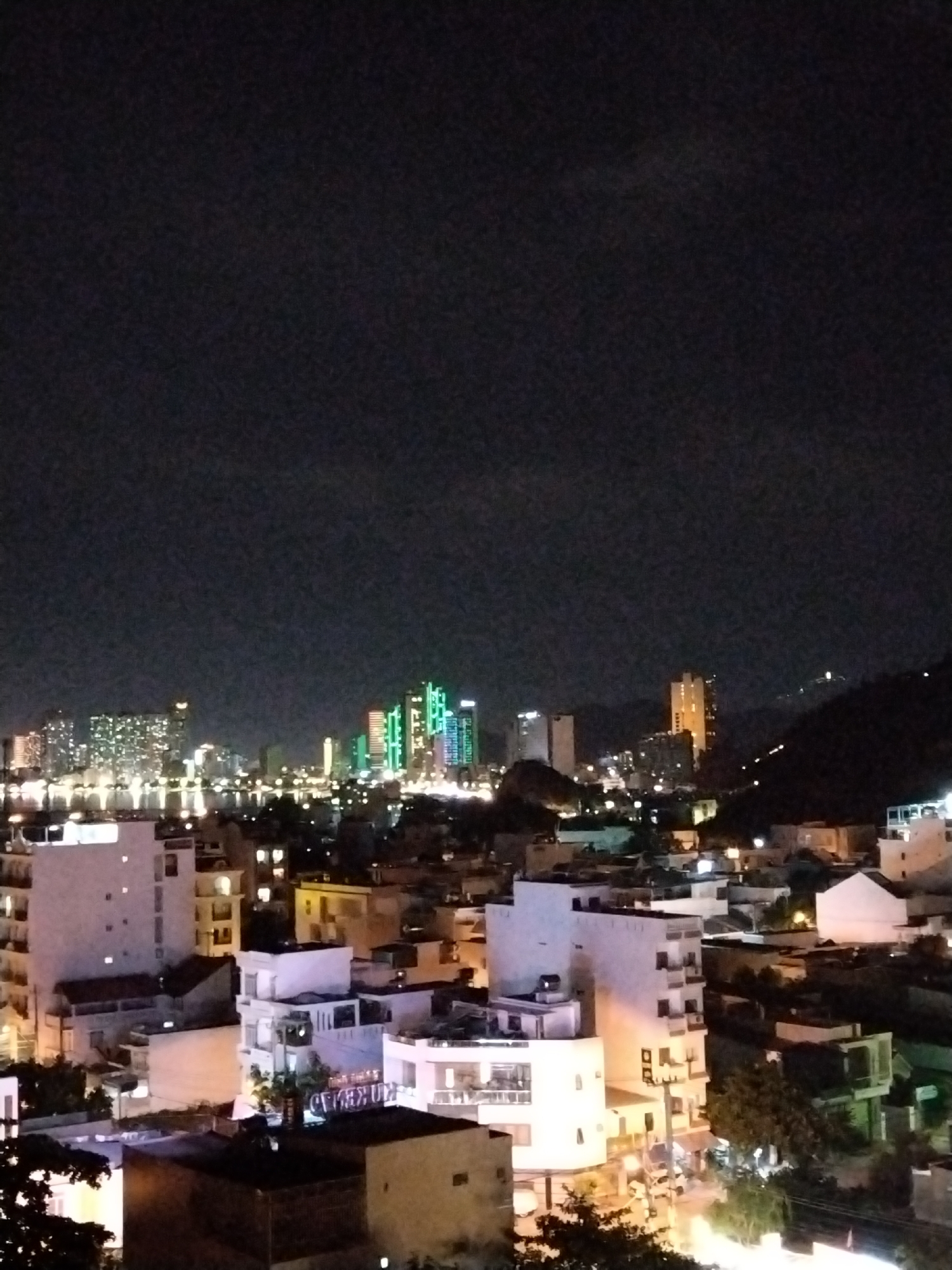This is a nighttime, elevated, horizontal, full-shot photograph of the Bac Nha Trang Ward in Vietnam. The scene is dominated by the dark night sky, with a cluster of illuminated buildings in the midground and foreground. In the distance, several high-rise buildings are visible, some with brightly colored lights, including green and blue. Closer to the foreground, there are lower-rise buildings with illuminated windows and balconies. Some of the buildings in the foreground are white, while others appear to be made of concrete. Trees are visible in the lower part of the image, with some illuminated by artificial light. The overall impression is of a lively urban environment at night. There are no people or animals visible in the frame.