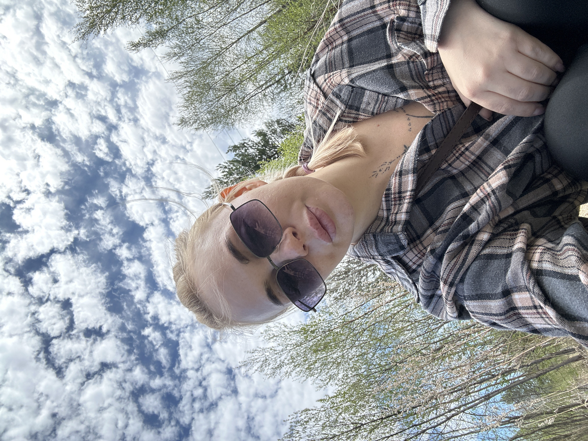 A woman wearing sunglasses and a plaid shirt looks down at the camera. A tattoo is visible on her neck. She is outside, with trees and a cloudy sky visible in the background. The location cannot be determined as Высокий, Russia, based on the image.