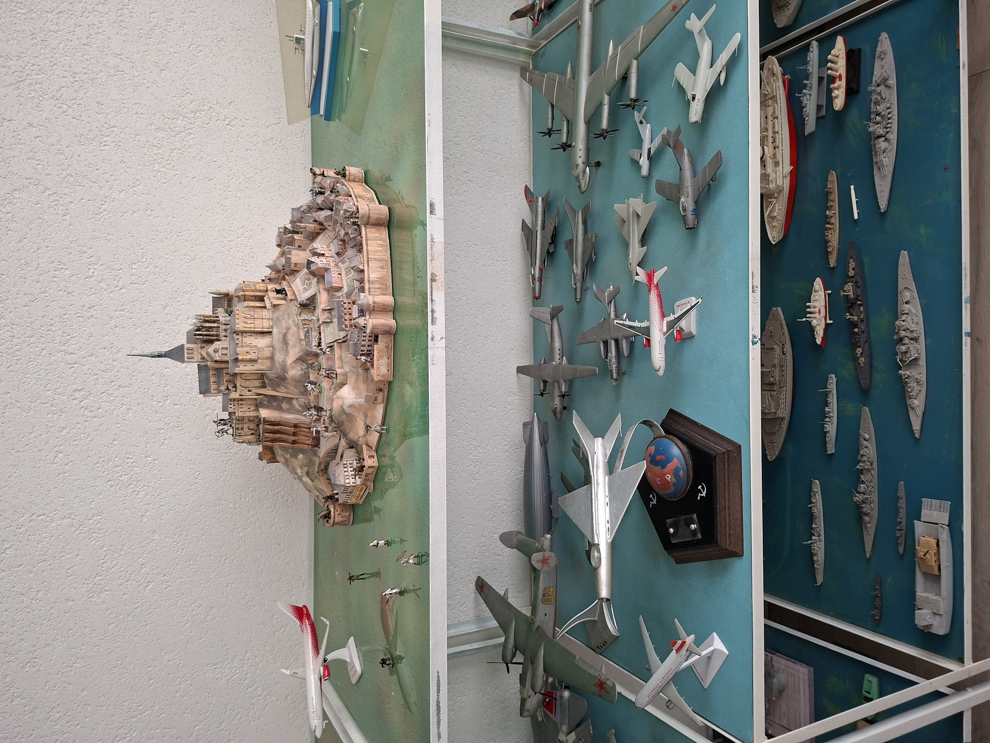 The image displays a collection of model vehicles and structures arranged on shelves. The top shelf features a diorama of a fortified medieval town built on a rocky island, resembling Mont Saint-Michel. Small figures of people are scattered throughout the diorama. To the left of the diorama is a model airplane. To the right, a model of a submarine rests on a blue base.
The middle shelf is filled with various model airplanes and a zeppelin. These models are predominantly grey and silver, with some exhibiting red star markings, suggesting a military theme.
The bottom shelf showcases a collection of model ships and boats on a blue surface. These include what appear to be warships, a large passenger liner, and smaller vessels. There is no indication of the city of Ialoveni, Moldova in the image.