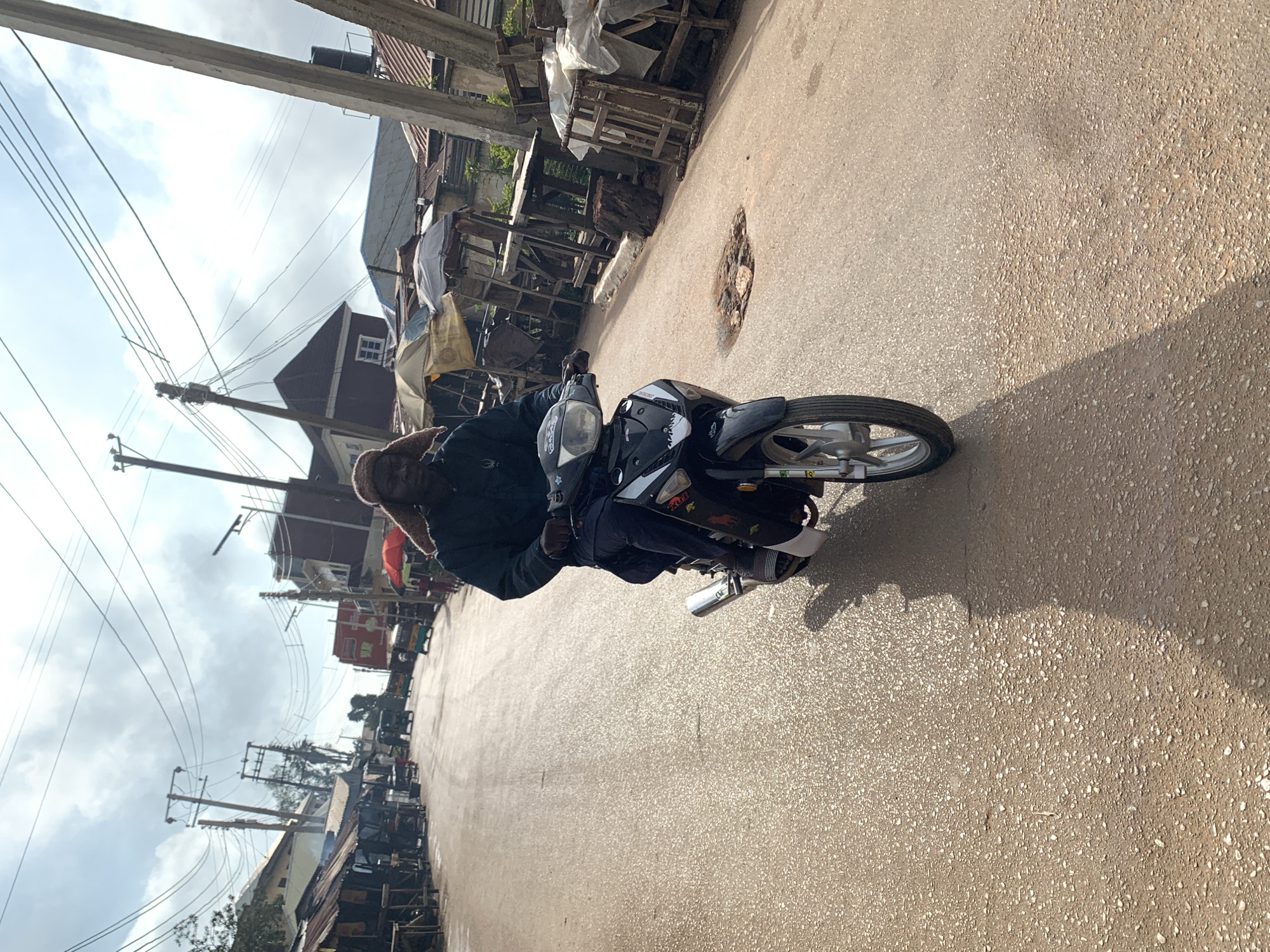 The image shows a street scene in Jos, Nigeria. A man is riding a black motorcycle down a paved road. He is wearing a dark jacket and a brown hat. The road is lined with various stalls and buildings, some with corrugated metal roofs. There are also utility poles and wires visible in the background, as well as a few other vehicles, including what appear to be auto-rickshaws. The sky is overcast with scattered clouds. The road surface appears to be asphalt with scattered aggregate and some patches of wear, including a noticeable pothole. The lighting suggests it is daytime, with sunlight casting shadows.