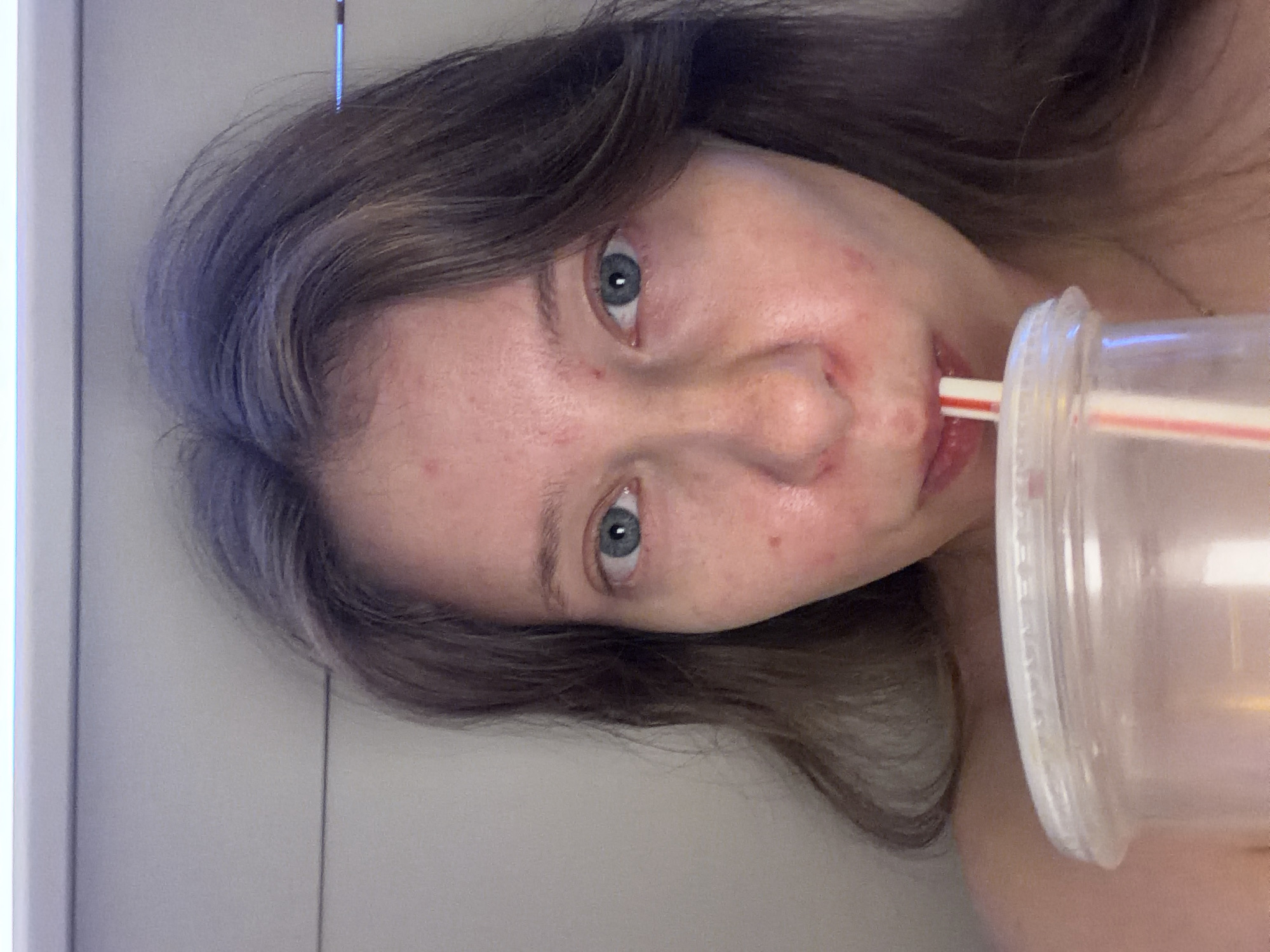 A woman is captured in a close-up selfie, holding a clear plastic cup with a red and white striped straw, from which she is drinking. Her head is tilted slightly, and her gaze is directed towards the camera. The background appears to be a wall with horizontal lines, possibly in a home or indoor setting. No specific city or location in Portland, United States of America is identifiable from the image. The lighting is even, suggesting it may be artificial.