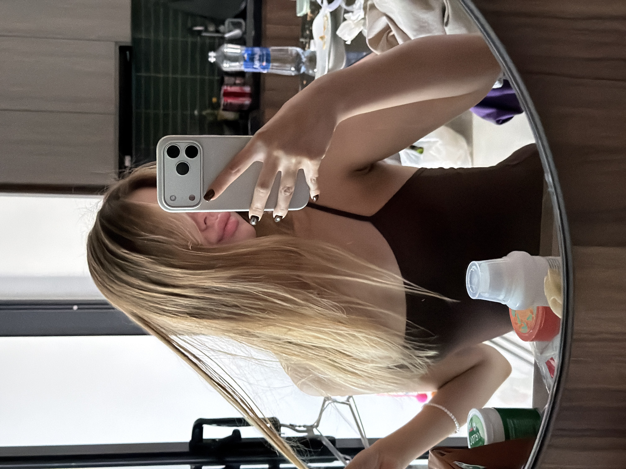 A young woman with blonde hair is taking a mirror selfie in what appears to be a bathroom or kitchen area. She is wearing a dark brown tank top and has on dark nail polish. Her hair is falling over her face as she holds up a silver smartphone with her right hand. In the background, there is a bottle of water, some containers, and what looks like a towel or clothing. The overall environment suggests a domestic setting, possibly an apartment or hotel room. Without specific landmarks or context, it is impossible to determine if this location is in Da Nang, Vietnam.