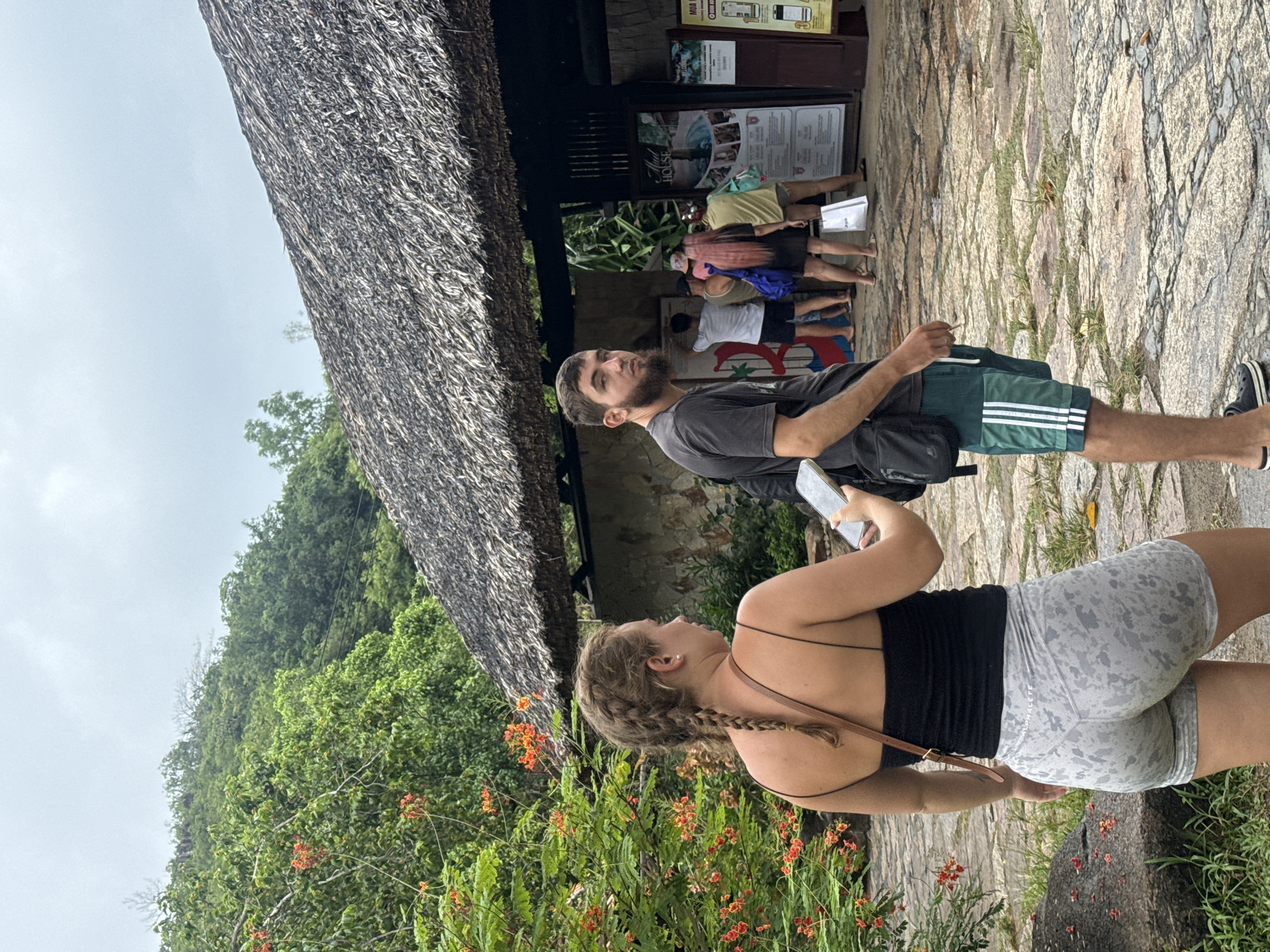 In Nha Trang, Vietnam, an overcast day frames a scene with two individuals in the foreground. A woman with braided hair, wearing a black tank top and grey patterned shorts, looks towards her right while holding a phone. To her right, a man with a beard and dark t-shirt, wearing green athletic shorts with white stripes, faces the viewer. Behind them, other visitors are visible near a thatched-roof building, suggesting an entrance or exhibit area. Lush green foliage and orange flowers surround the paved walkway, hinting at a tropical or natural park setting.