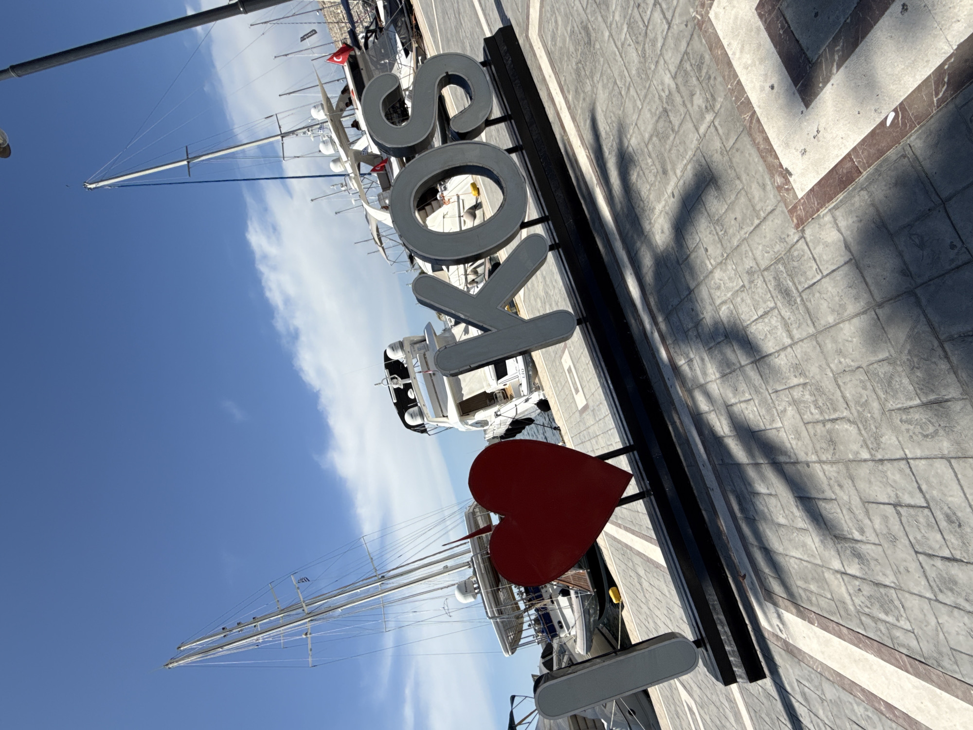 The photograph depicts the "I Love Kos" sign in the harbor of Kos, Greece. The sign, rendered in gray lettering, stands perpendicular to the paved walkway. A red heart replaces the word "Love." Behind the sign, sailboats are docked in the harbor, their masts reaching towards a blue sky with scattered clouds. The walkway is paved with gray stone, marked with a dark shadow cast by the sign.