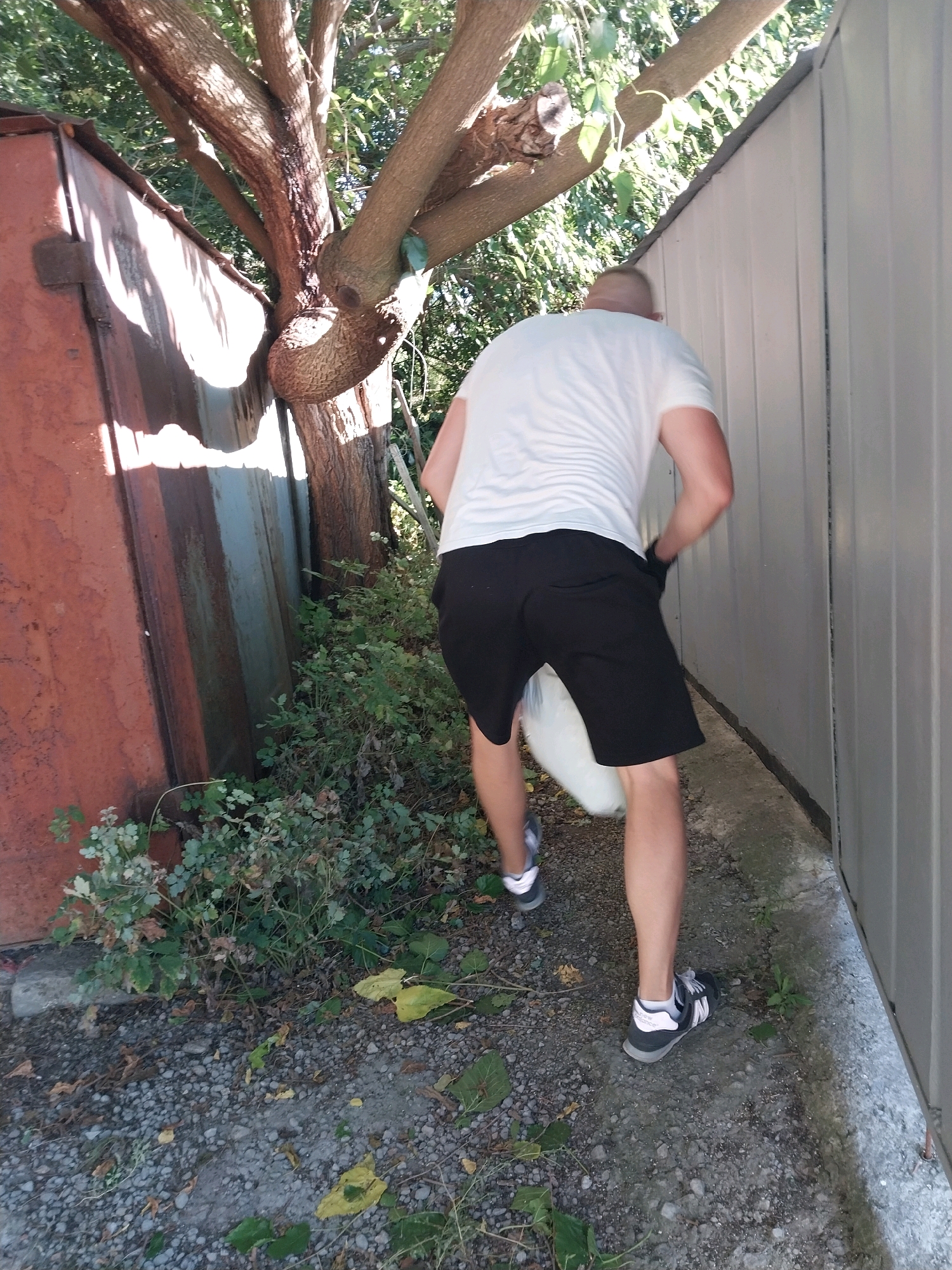 A bald man wearing a white t-shirt and black shorts is standing in a narrow alleyway, partially obscured by a tree and dense foliage. He is facing away from the camera, towards a light-colored, corrugated metal wall. In his right hand, he appears to be holding or carrying a white, bulky object. His left hand is in his pocket. He is wearing black and white athletic shoes. To his left is a rusty, reddish-brown corrugated metal structure. The ground is covered with gravel, dirt, and scattered leaves. The scene appears to be outdoors, possibly in a backyard or an industrial area. The location is identified as Dnipro, Ukraine.