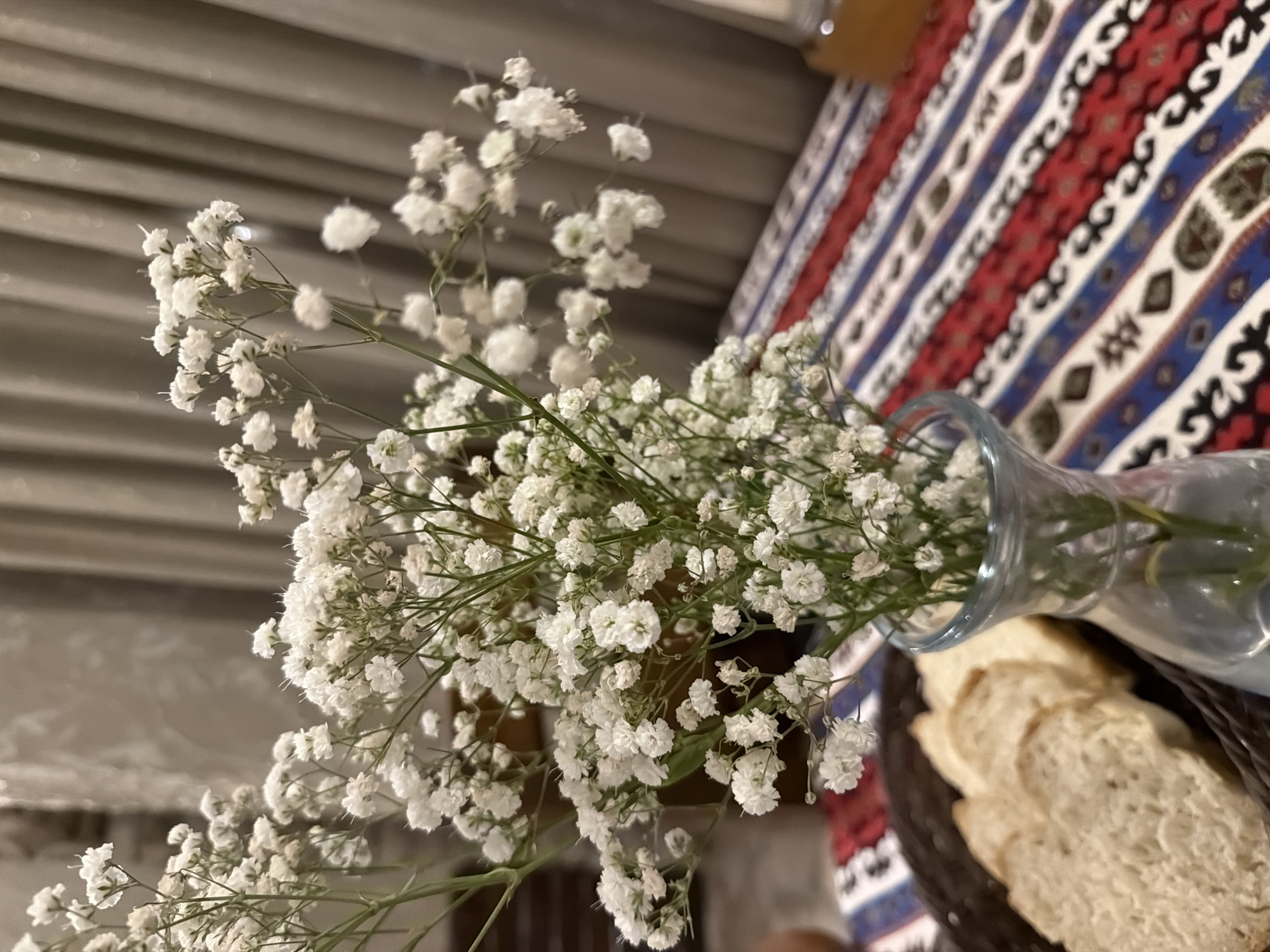 In Kumbor, Montenegro, a close-up shot captures a delicate arrangement of white baby's breath flowers in a clear glass vase. The vase sits on a table adorned with a vibrantly patterned tablecloth featuring red, blue, and white stripes with intricate ethnic motifs. To the left of the vase, a dark woven basket holds several slices of bread, suggesting a meal setting. In the background, neutral-toned curtains hang, creating a soft, diffused light that highlights the floral display and the textures of the table setting. The overall scene evokes a sense of simple elegance and domestic tranquility.