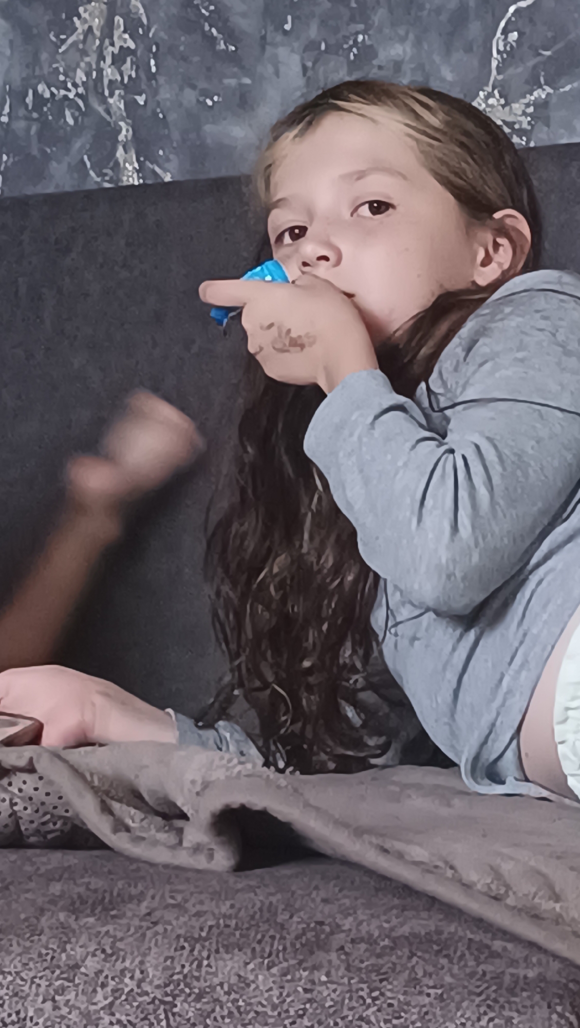 The image depicts a young person lying on a couch, with a blurry hand visible in the background. The person in the foreground is wearing a grey hoodie and has long, dark, curly hair with lighter streaks. They are holding a small, blue object to their mouth. The couch appears to be dark grey. The background wall has a textured, grey pattern. The overall setting suggests an indoor, casual environment. There is no information available regarding the location in the city of Karsha, Russia.