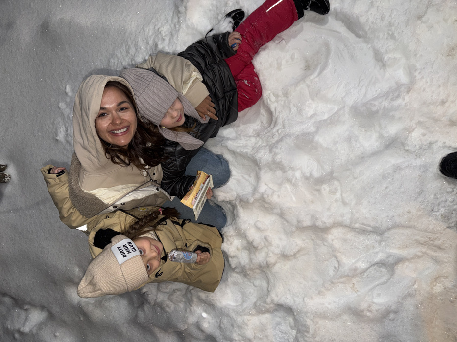 Three individuals are depicted outdoors in deep, undisturbed white snow, illuminated under nighttime or low-light conditions in Krasnoyarsk, Russia.
An adult female is positioned centrally, exhibiting a broad smile directed towards the camera. She wears a light beige hooded winter jacket and has dark brown hair. Her left arm is extended around a child seated to her right. This child, appearing female, wears a grey knitted winter hat covering the ears, a dark padded jacket, and bright red snow pants. The child holds a small, foil-wrapped rectangular item in the right hand.
To the adult's left, another child, appearing female, is present. This child wears a beige knitted beanie with a white rectangular patch bearing the text "DIRTY HAIR CLUB," a tan winter coat, and light blue denim trousers. The child holds a clear, cylindrical container in the right hand and smiles towards the camera.
All subjects are dressed in cold-weather garments. The snow exhibits natural undulations and texture across the entire visible ground. A portion of a dark shoe is visible at the bottom edge of the image.