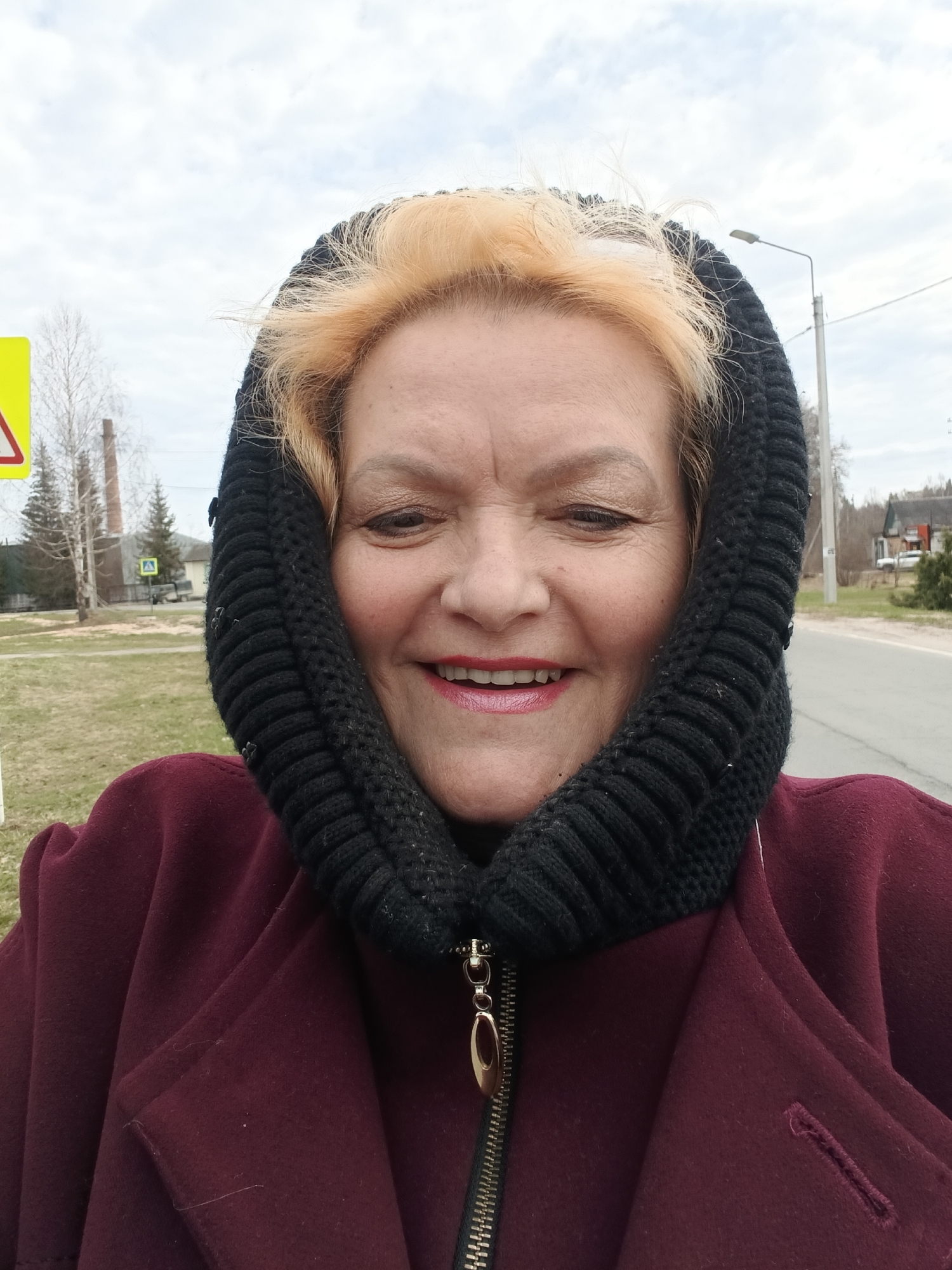 A close-up selfie photograph shows a smiling middle-aged woman in an outdoor urban setting. She is wearing a maroon coat with a partially zipped front, and a black knitted hood frames her face. Her hair is blonde. A road runs to the right side of the photo, with houses and vegetation visible in the background. On the left, there is a yellow road sign and bare trees. The sky is overcast. Based on landmarks, this photograph was taken in Красный Маяк, Russia.