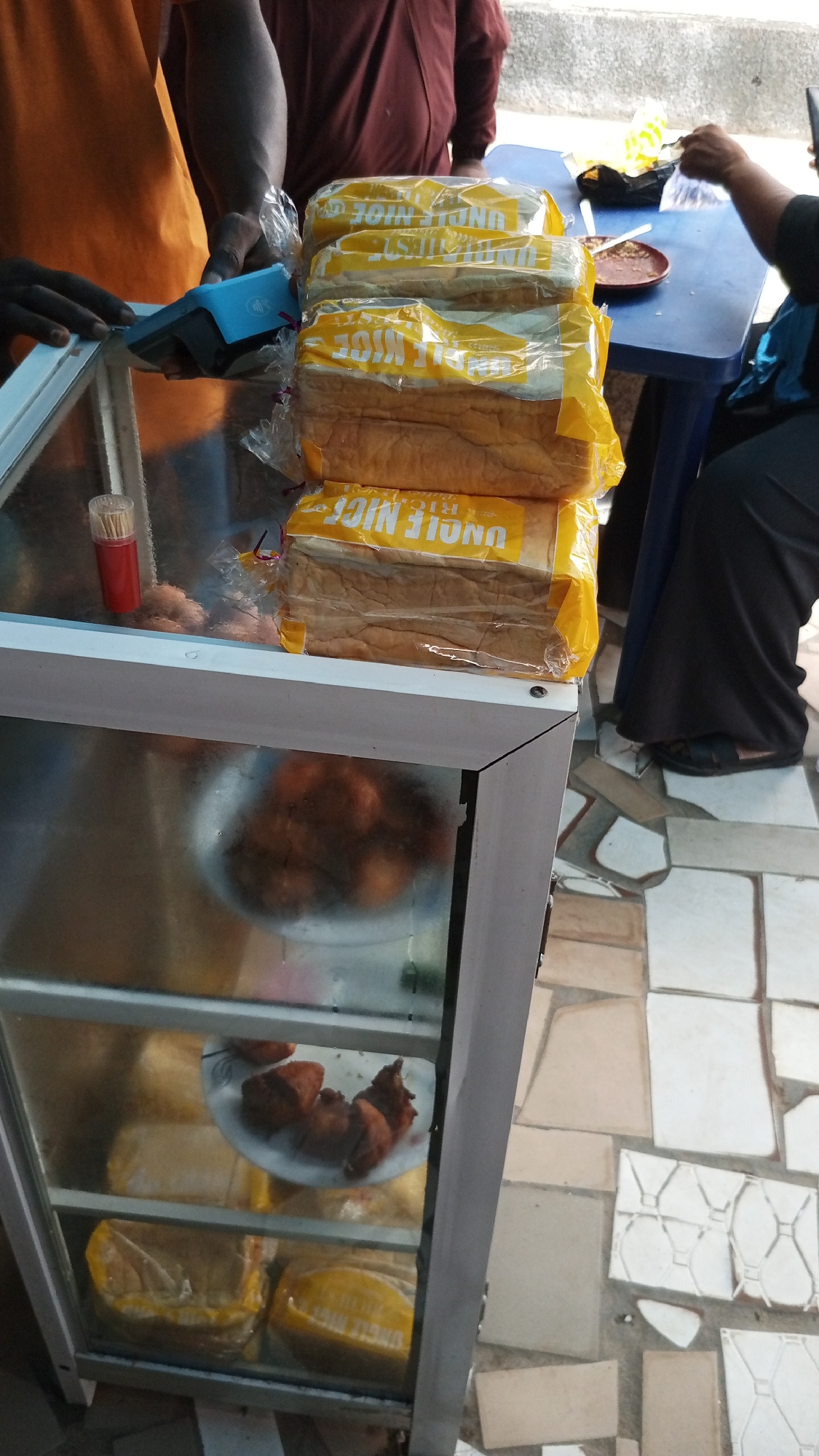 This image depicts a street food vendor in Lokoja, Nigeria, showcasing a variety of items. The central focus is a metal-framed glass display case containing several plates of fried snacks and loaves of bread in yellow packaging labeled "UNCLE NICE." A stack of these bread loaves sits atop the display case. A person in an orange shirt stands next to the case, operating a blue Point-of-Sale (POS) device. To the right, a blue plastic table is visible with a person seated, appearing to be eating from a plate. The tiled pavement suggests an outdoor setting.