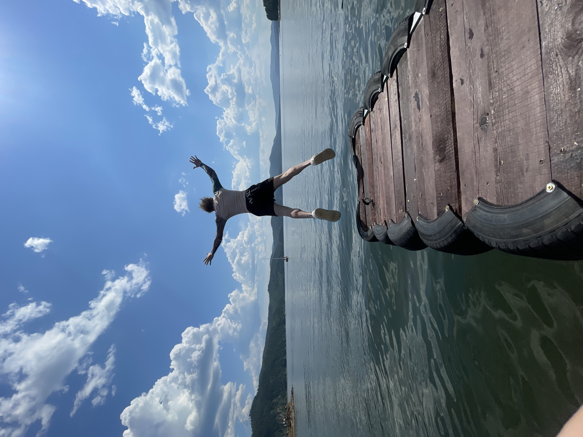 A person is captured mid-air, jumping from a wooden dock into a body of water. The image is oriented vertically, with the sky taking up a significant portion of the frame on the left. The person, seen from behind, is wearing black shorts and a light-colored t-shirt, with their arms and legs outstretched as if in a dive. They have tattoos on their left arm.
The dock, made of weathered wooden planks, occupies the right side of the image. It appears to be reinforced with sections of tires along its edge. The water is calm, reflecting the bright blue sky and fluffy white clouds. In the distance, a tree-covered hillside stretches across the horizon, with a hazy mountain range beyond. A small sailboat can be seen on the water.
The weather is sunny with scattered clouds, suggesting a clear, pleasant day. The overall mood is one of exhilaration and freedom associated with summer activities and water sports. The scene is likely set during daylight hours, given the brightness and shadows. The context provided places the location in Divnogorsk, Russia. No visible text is present in the image.