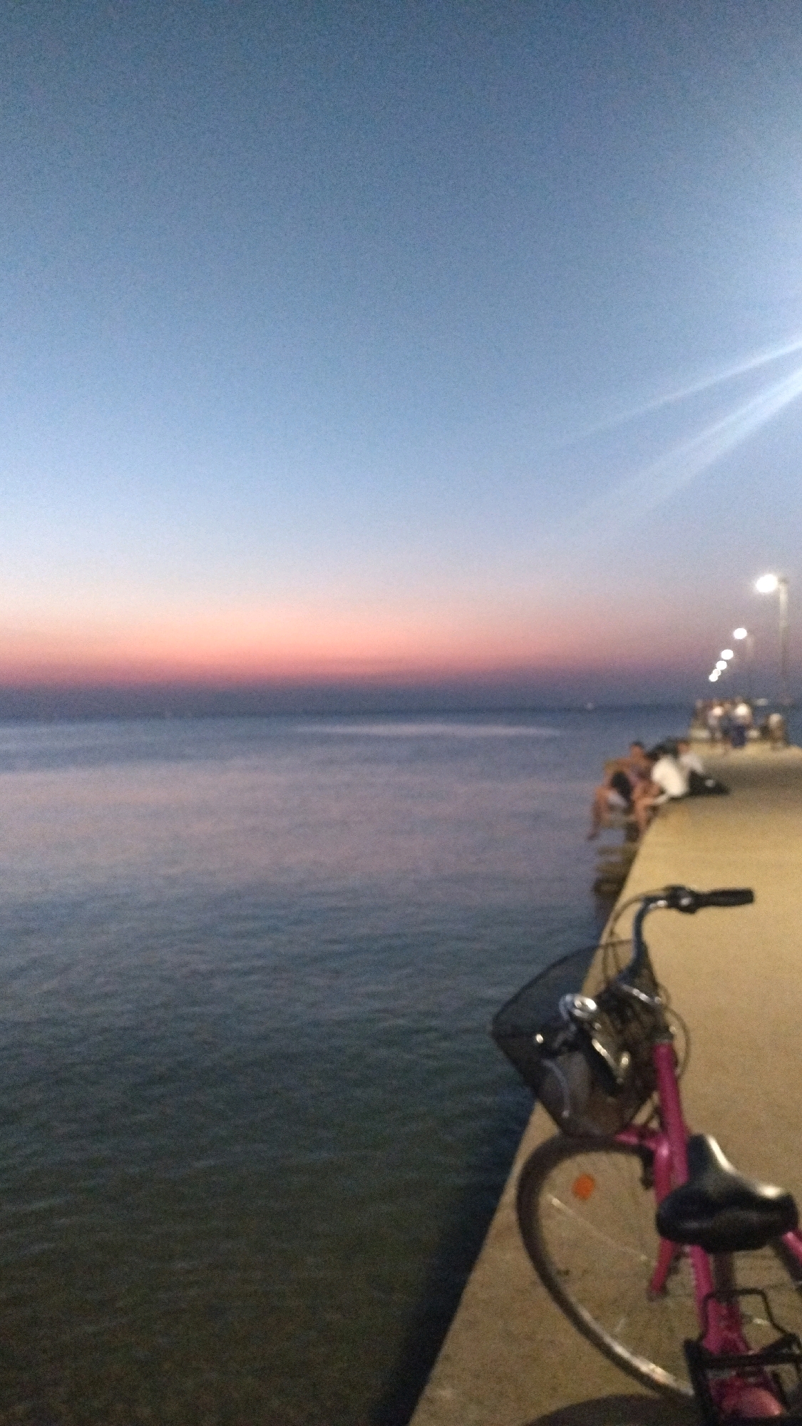 The image depicts a waterfront scene at dusk, likely in Perea, Greece, with a pink bicycle in the foreground. The sky transitions from a pale blue at the top to soft pink and orange hues near the horizon, indicating sunset or sunrise. The sea is calm and stretches to the horizon, meeting the colorful sky.
A concrete pier extends into the water from the right side of the frame, lined with lampposts that cast a warm glow. Several people are visible on the pier, some sitting with their legs dangling over the edge and others standing. They appear to be casually observing the sunset over the water. The pink bicycle, parked on the pier, has a basket on the front and a rear rack. The overall atmosphere is serene and recreational, capturing a moment of twilight enjoyment by the sea.
