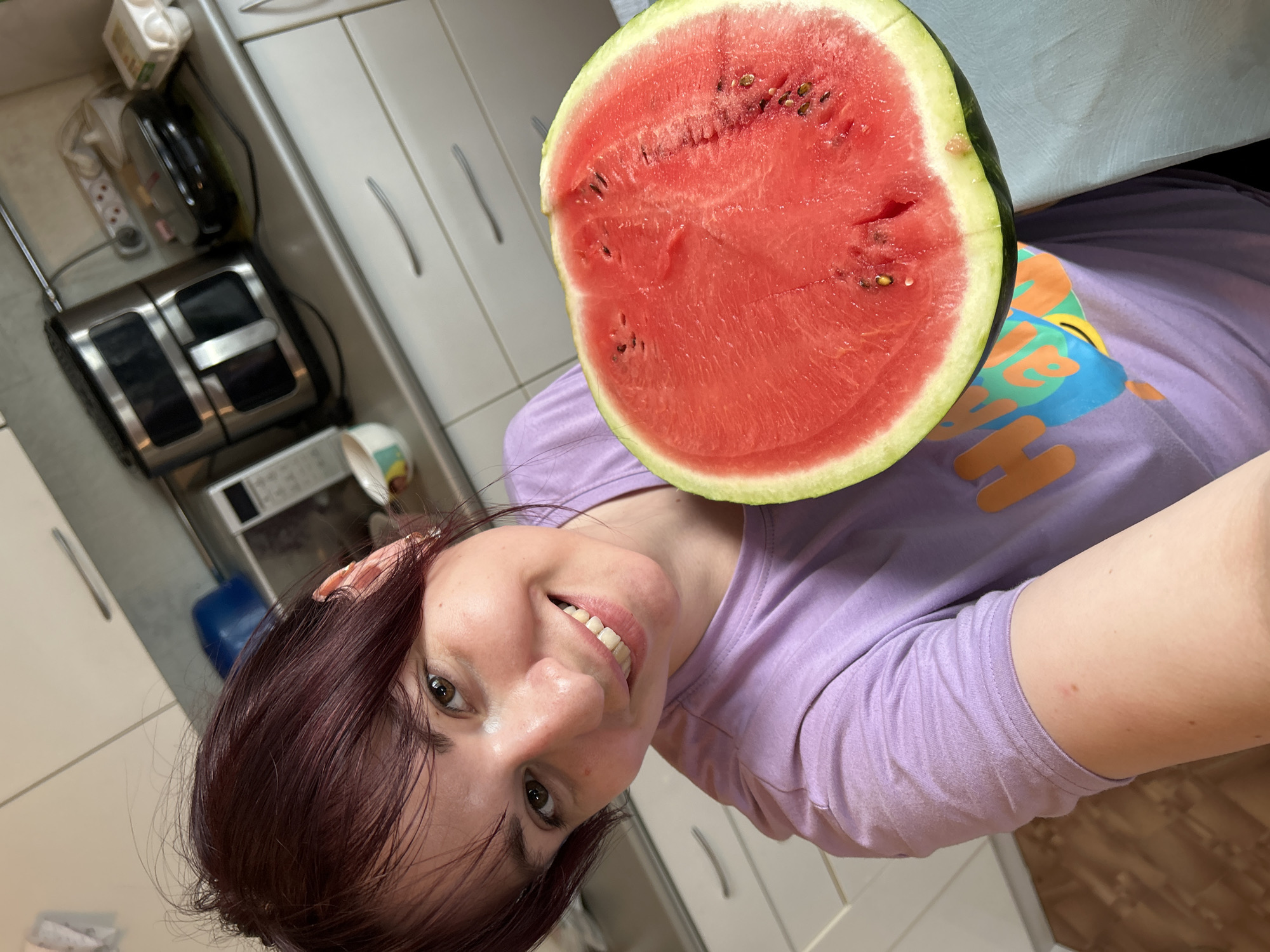 A woman with short, dark red hair, wearing a lavender t-shirt, smiles at the camera while holding a slice of watermelon. The watermelon is a vibrant red with visible seeds and a green rind. She is in a kitchen, with white cabinets and countertops visible in the background. Appliances such as a microwave and an air fryer are also present. The overall setting appears to be residential. The location in the city of Фоки, Russia is not determinable from the image.